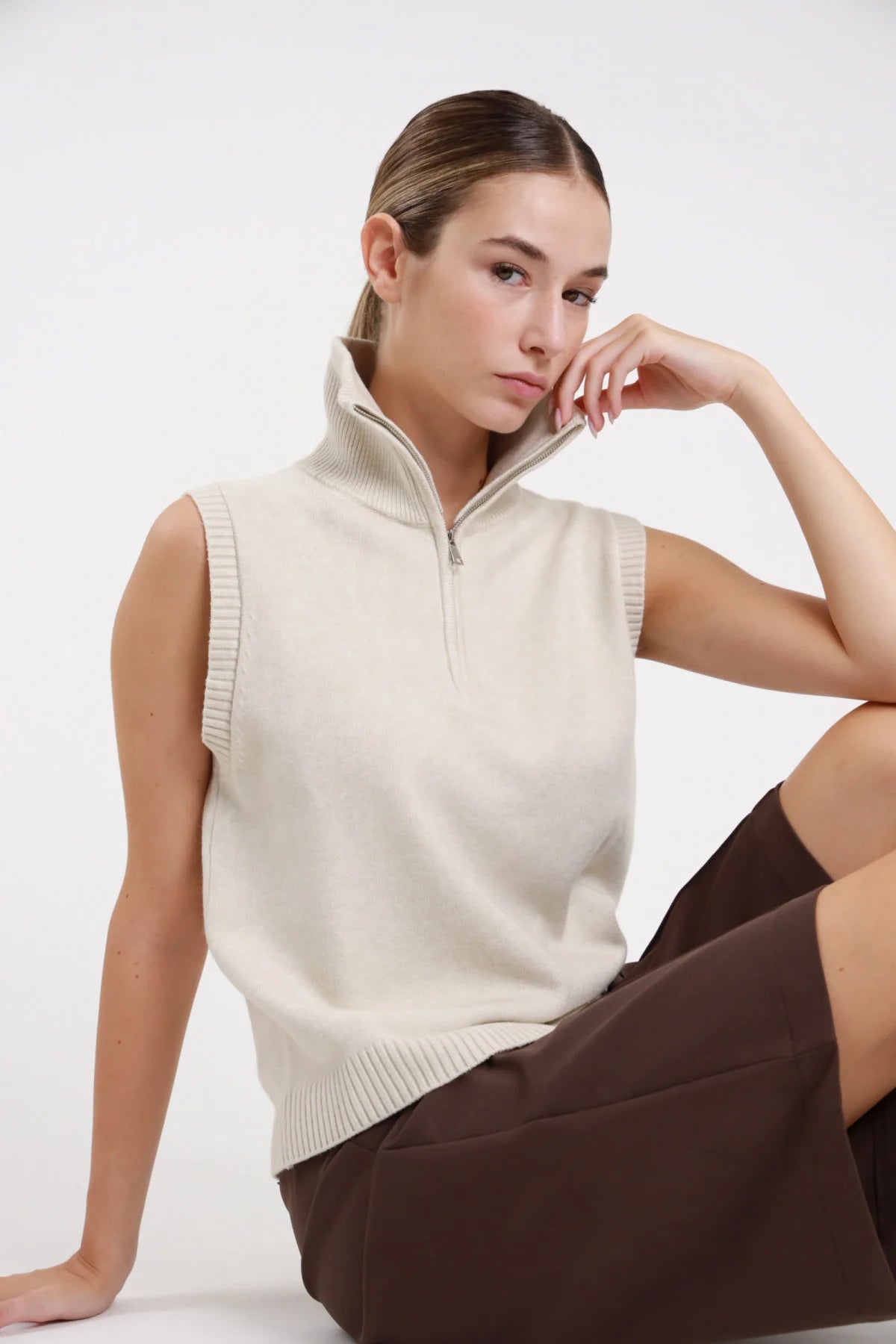 Gilet zip maglia collo alto colore panna - modella seduta posa laterale mano al mento shorts marroni capelli raccolti