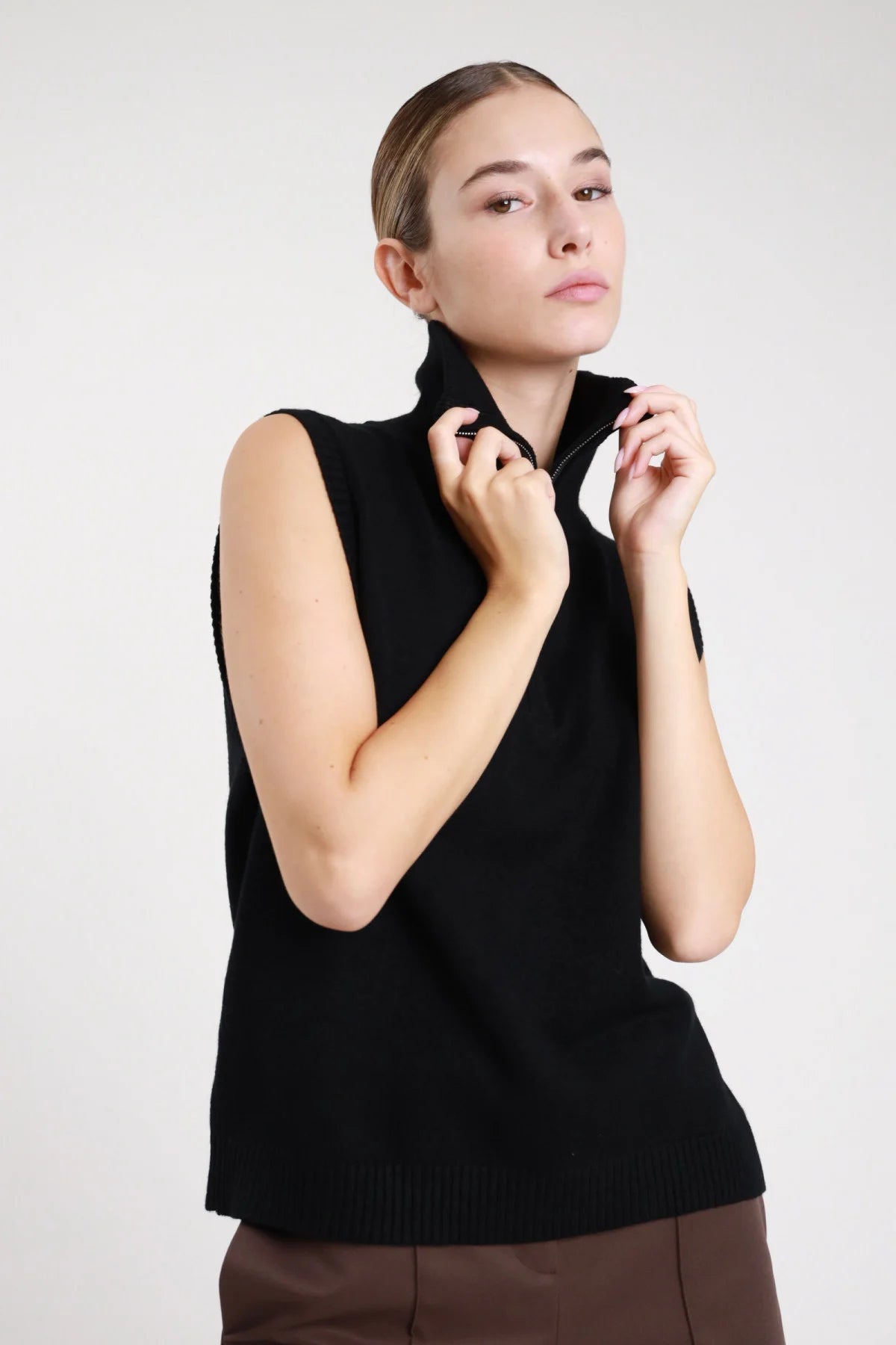 Gilet zip maglia nero collo alto - modella busto frontale mani sulla zip sguardo laterale pantaloni marroni capelli raccolti