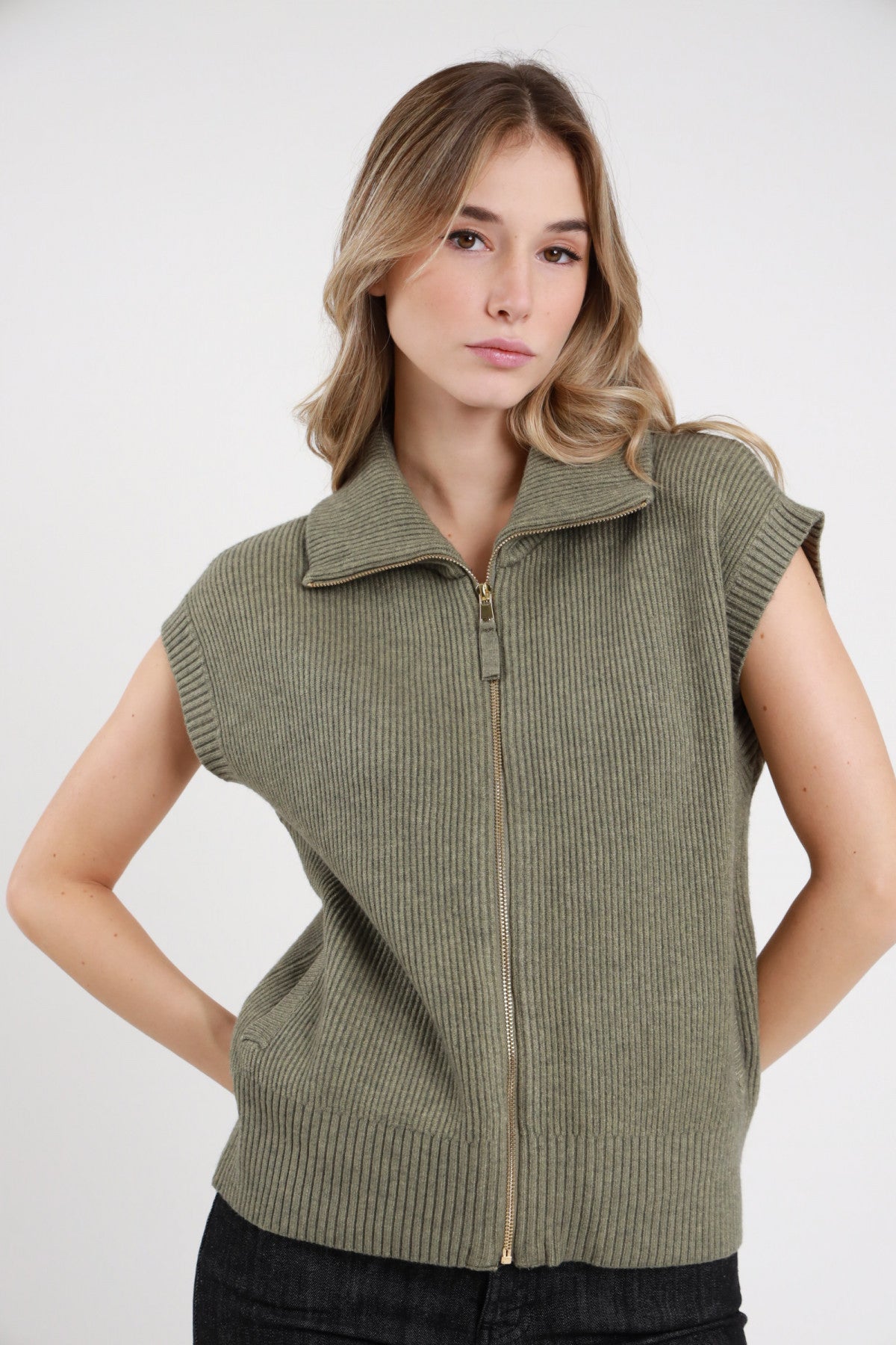 Gilet Costa Inglese Zip verde militare oliva 50% viscosa costa inglese costine larghe verticali colletto polo alto costina zip dorata frontale cursore grande cap sleeve costina bordo elasticizzato silhouette boxy frontale busto mani fianchi - modella busto frontale mani fianchi sguardo frontale capelli biondi mossi sfondo bianco jeans nero slim abbinati