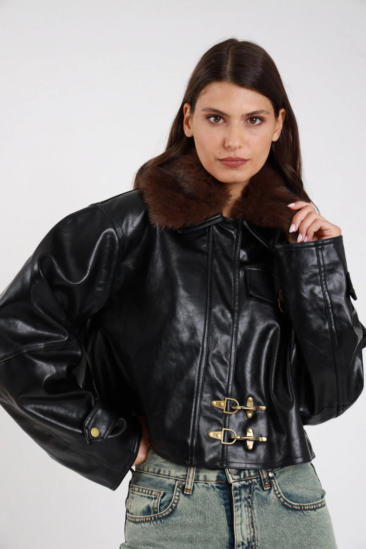 Giacca ecopelle lucida nero colletto ecopelliccia marrone amovibile fibbie ganci dorate balloon oversize boxy crop frontale busto mano colletto - modella frontale busto mano colletto ecopelliccia sguardo frontale capelli castani scuri ondulati sfondo bianco jeans grigio chiaro