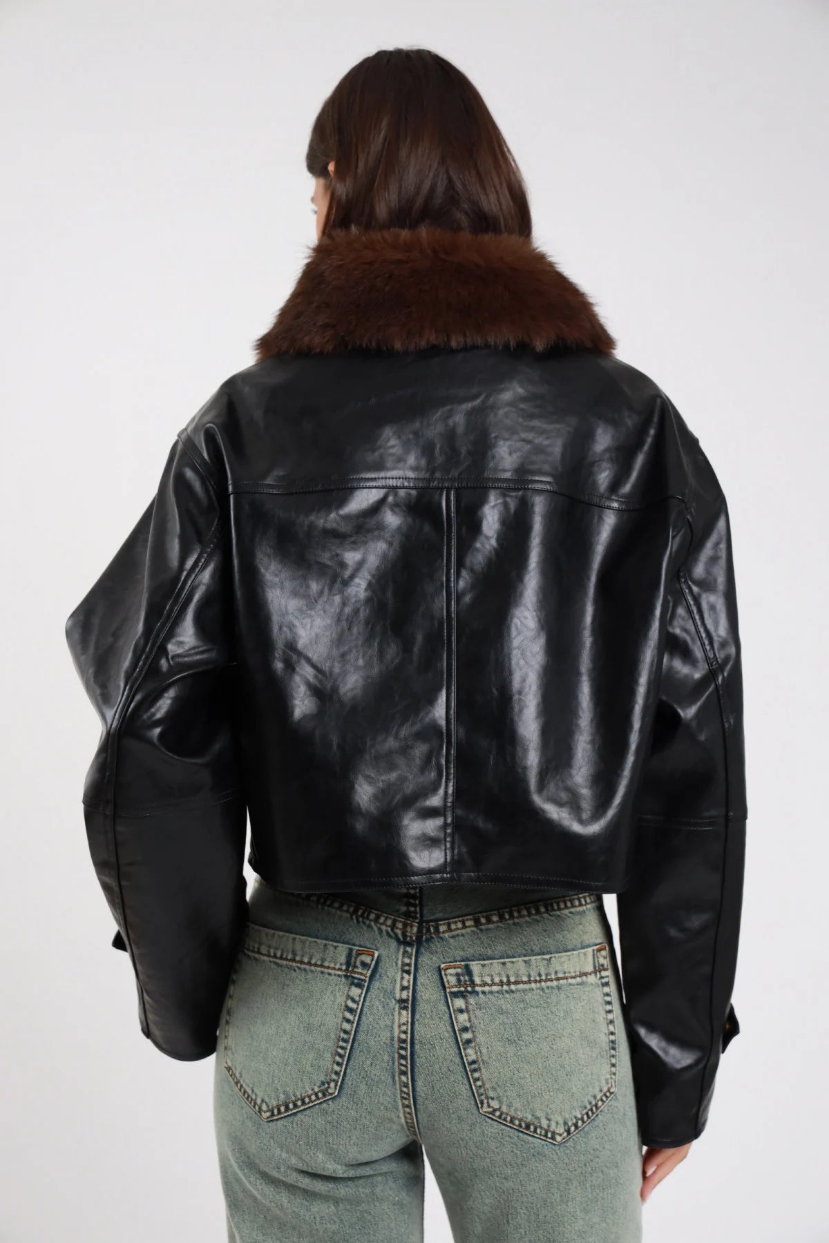 Giacca ecopelle lucida nero colletto ecopelliccia marrone amovibile fibbie ganci dorate balloon oversize boxy crop retro intera - modella retro intera sguardo non visibile capelli castani scuri sfondo bianco jeans grigio chiaro