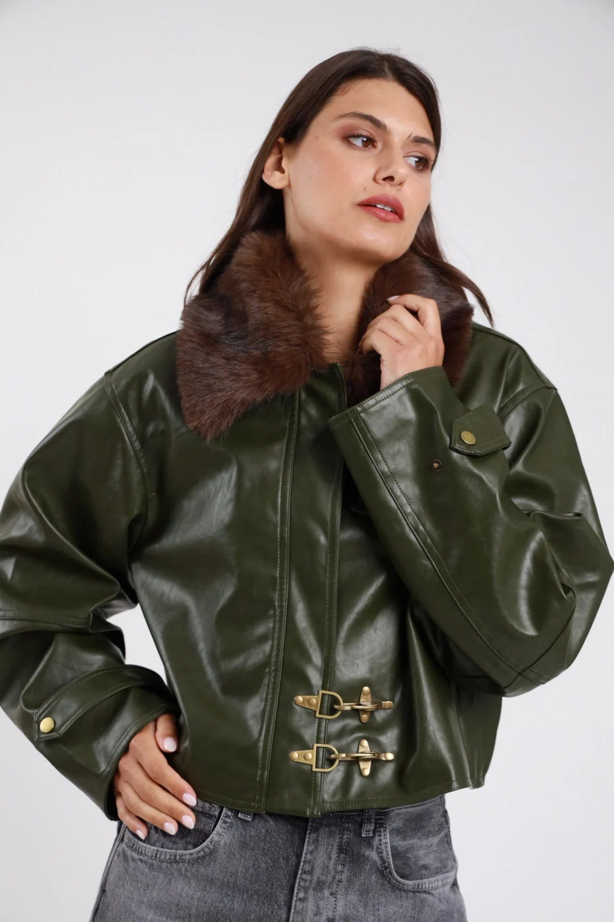 Giacca ecopelle lucida verde militare khaki colletto ecopelliccia marrone amovibile fibbie ganci dorate balloon oversize boxy crop frontale busto mano colletto - modella frontale busto mano colletto ecopelliccia sguardo laterale capelli castani scuri ondulati sfondo bianco jeans grigio chiaro