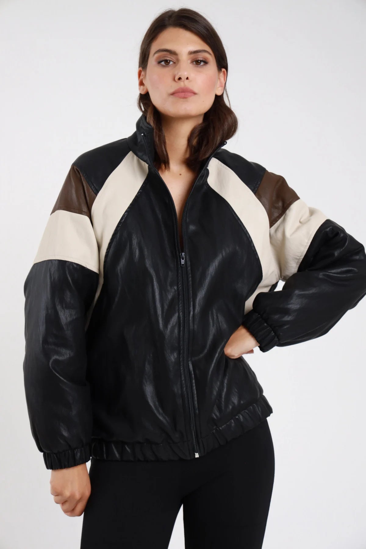 Giacca ecopelle bomber oversize nero donna inserti color-block panna marrone spalle maniche collo alto zip frontale fascia costine - vista frontale intera giacca aperta mano sul fianco con leggings neri