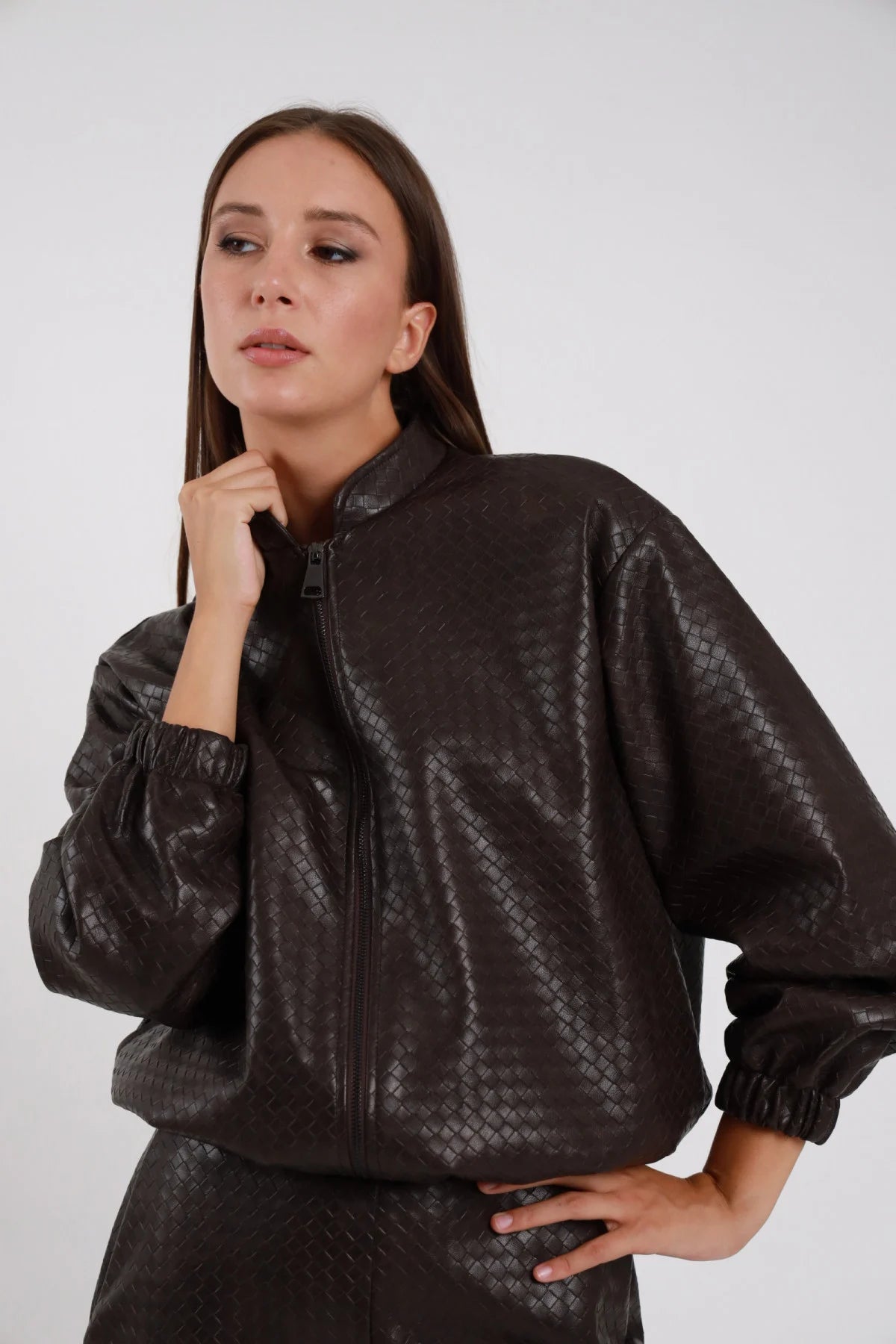 Bomber Ecopelle Intrecciato marrone cioccolato scuro woven embossed romboidale oversize boxy crop zip chiusa tono su tono mano mento colletto stand-up manica balloon polsino elastico arricciato busto frontale 3/4 mano fianchi - modella busto frontale 3/4 mano mento sguardo laterale capelli castani lisci sfondo bianco gonna coordinata marrone