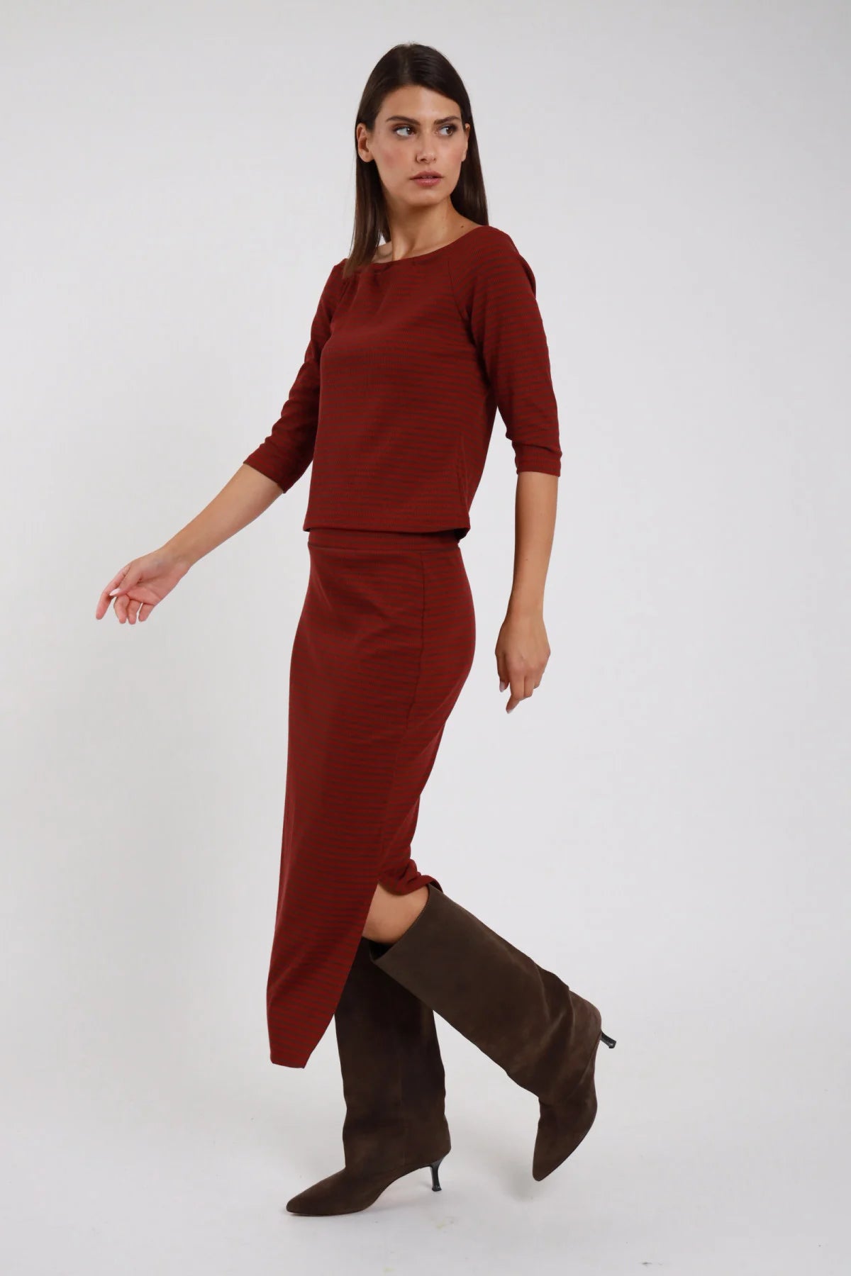 Gonna Midi Longuette bordeaux mattone tinta unita 48% cotone elastico vita silhouette semi-aderente spacco laterale - modella intera laterale posa dinamica gamba avanzata sguardo laterale capelli castani lisci sfondo bianco top bordeaux coordinato stivali marrone ginocchio tacco