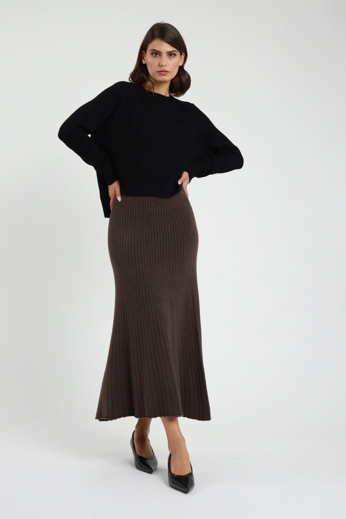 Gonna in Maglia coste ribbed marrone cioccolato vita alta silhouette sirena mermaid midi-maxi orlo svasato frontale intera - modella frontale intera mani fianchi sguardo frontale capelli castani sfondo bianco maglione oversize nero décolleté nere tacco