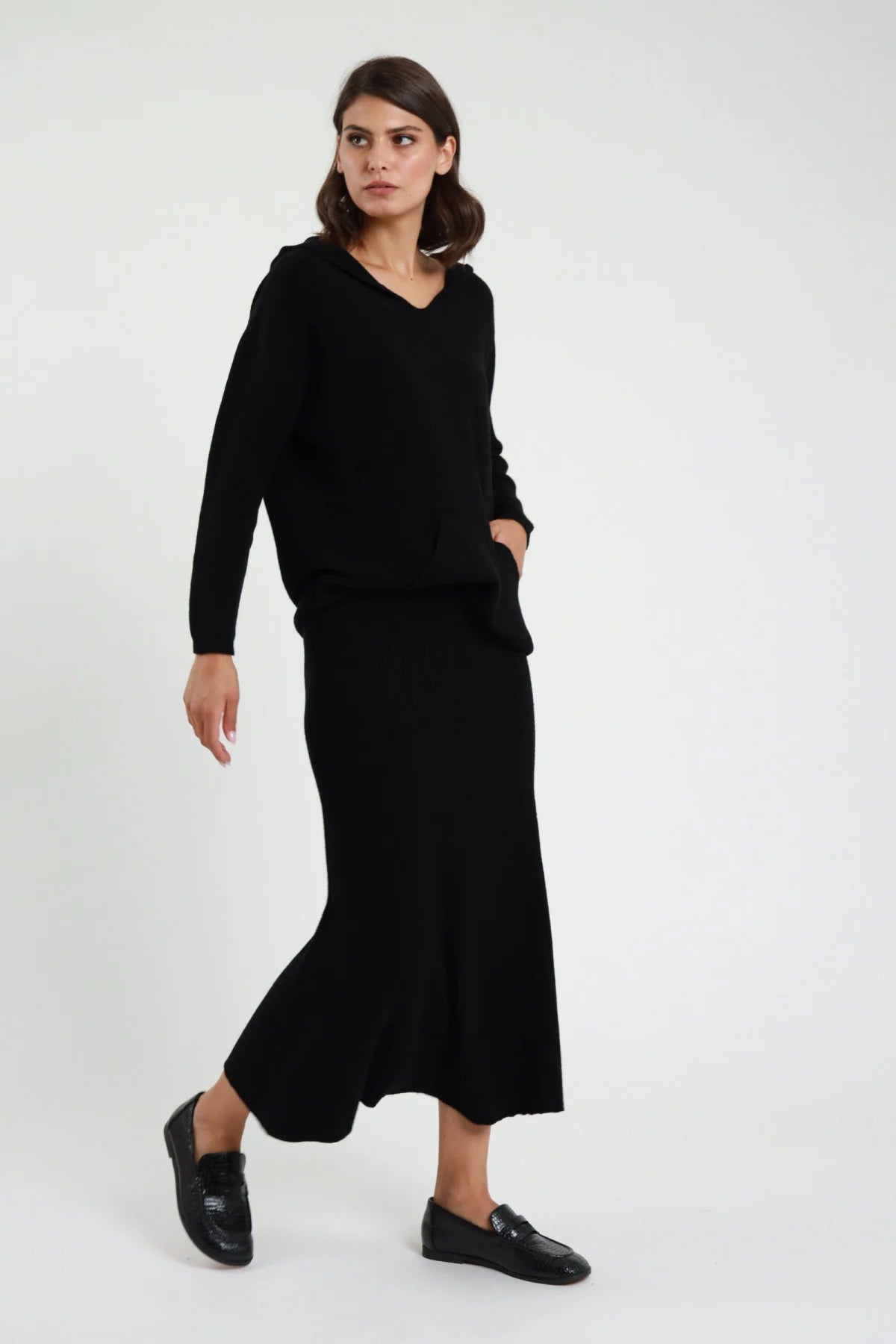Gonna in Maglia coste ribbed nero vita alta silhouette sirena mermaid midi-maxi orlo svasato frontale intera posa camminata - modella frontale intera posa camminata mano tasca sguardo laterale capelli castani sfondo bianco felpa maglia nera scollo V loafer neri vernice