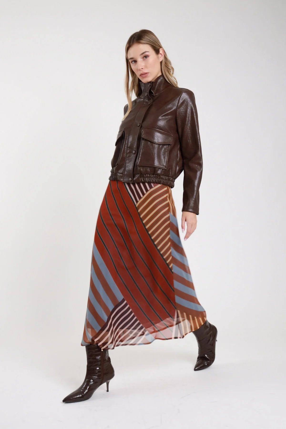 Gonna fantasia righe diagonali multicolore patchwork midi frontale intera giacca pelle marrone stivali croco tacco - modella frontale mano al fianco sguardo frontale capelli biondi