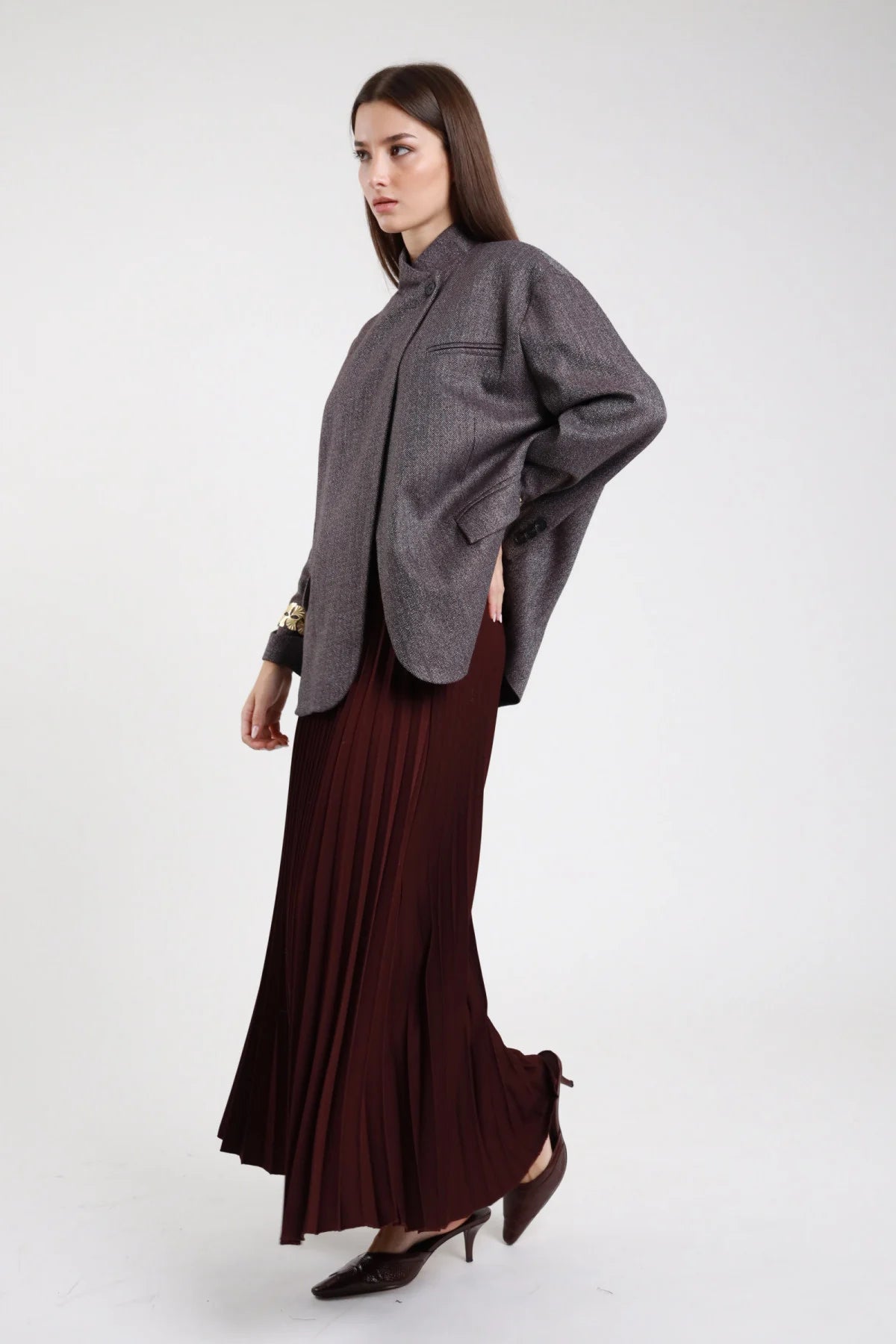 Gonna lunga plissé bordeaux prugna vinaccia laterale intera maxi floor-length vita alta plissé permanente allover corpo svasato fluente orlo dritto decolleté tacco scuri - modella laterale intera mano fianco sguardo frontale con blazer herringbone grigio spigato oversize bracciali oro decolleté tacco scuri capelli castani scuri lisci sfondo bianco