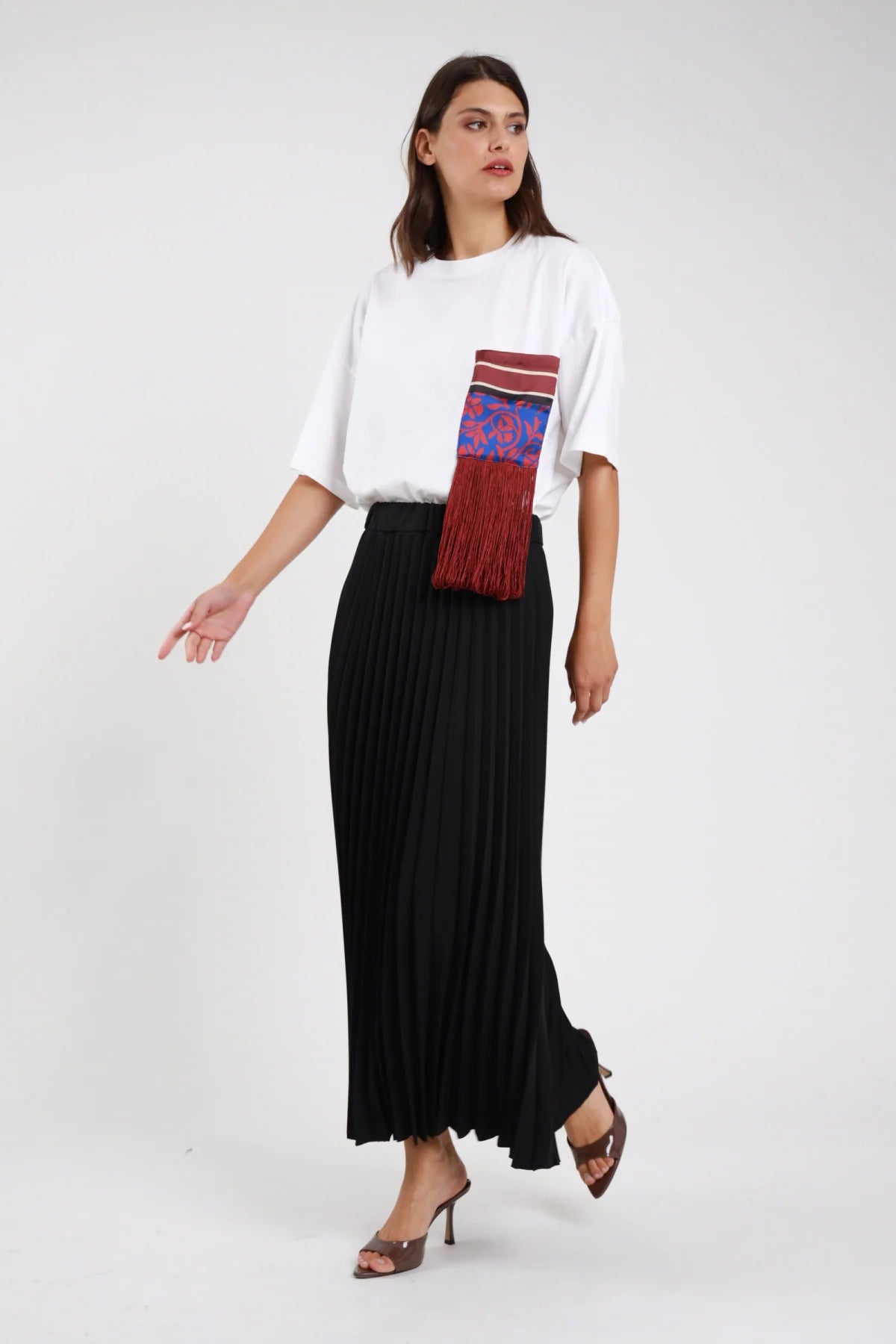 Gonna lunga plissé nero maxi floor-length vita alta elastico invisibile plissé fitto permanente allover corpo ampio svasato fluente orlo dritto sandali tacco trasparenti - modella frontale intera sguardo laterale con t-shirt taschina bianca stampa blu rosso frange bordeaux sandali tacco trasparenti marroni capelli castani scuri lisci sfondo bianco