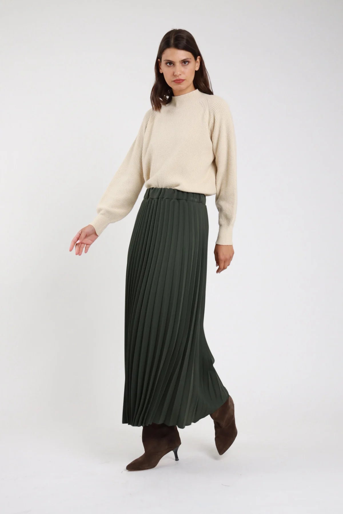 Gonna lunga plissé verde bottiglia foresta maxi floor-length vita alta elastico invisibile plissé fitto permanente allover corpo ampio svasato fluente orlo dritto stivali marroni tacco - modella frontale tre quarti dinamica passo sguardo laterale basso con pullover panna mock neck oversize stivali marroni tacco capelli castani scuri lisci sfondo bianco
