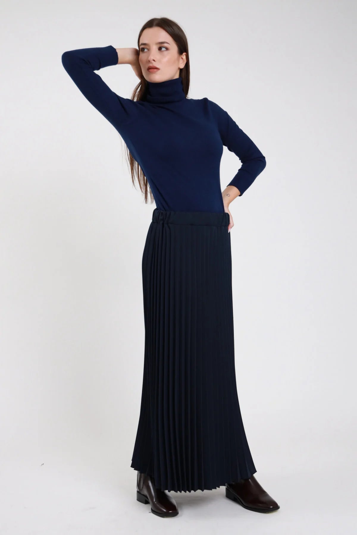 Gonna lunga plissé blu navy maxi floor-length vita alta elastico invisibile plissé fitto permanente allover corpo ampio svasato fluente orlo dritto ankle boots neri - modella frontale tre quarti mano nuca sguardo laterale con dolcevita blu navy aderente coordinato ankle boots neri capelli castani scuri lisci sfondo bianco