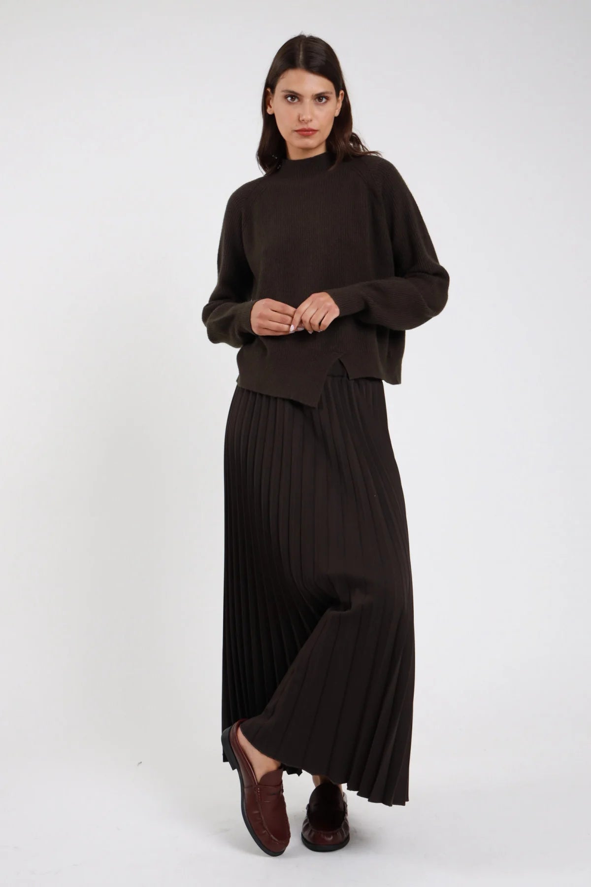 Gonna lunga plissé cioccolato moro scuro maxi floor-length vita alta elastico invisibile plissé fitto permanente allover corpo ampio svasato fluente orlo dritto loafer marroni - modella frontale intera mani incrociate sguardo frontale intenso con pullover mock neck moro coordinato loafer marroni capelli castani scuri lisci sfondo bianco