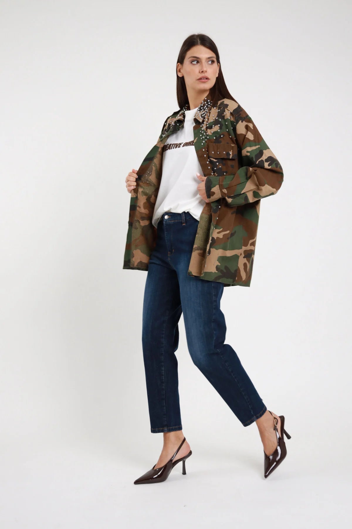 Jeans Chino Slim Vita Alta blu scuro indaco 98% cotone gamba dritta affusolata tasche frontali bottone argento fondo cropped caviglia lavaggio scuro indaco - modella intera 3/4 frontale posa camminata dinamica sguardo laterale alto capelli castani lisci sfondo bianco camicia militare mimetica aperta t-shirt bianca slingback marrone tacco spillo