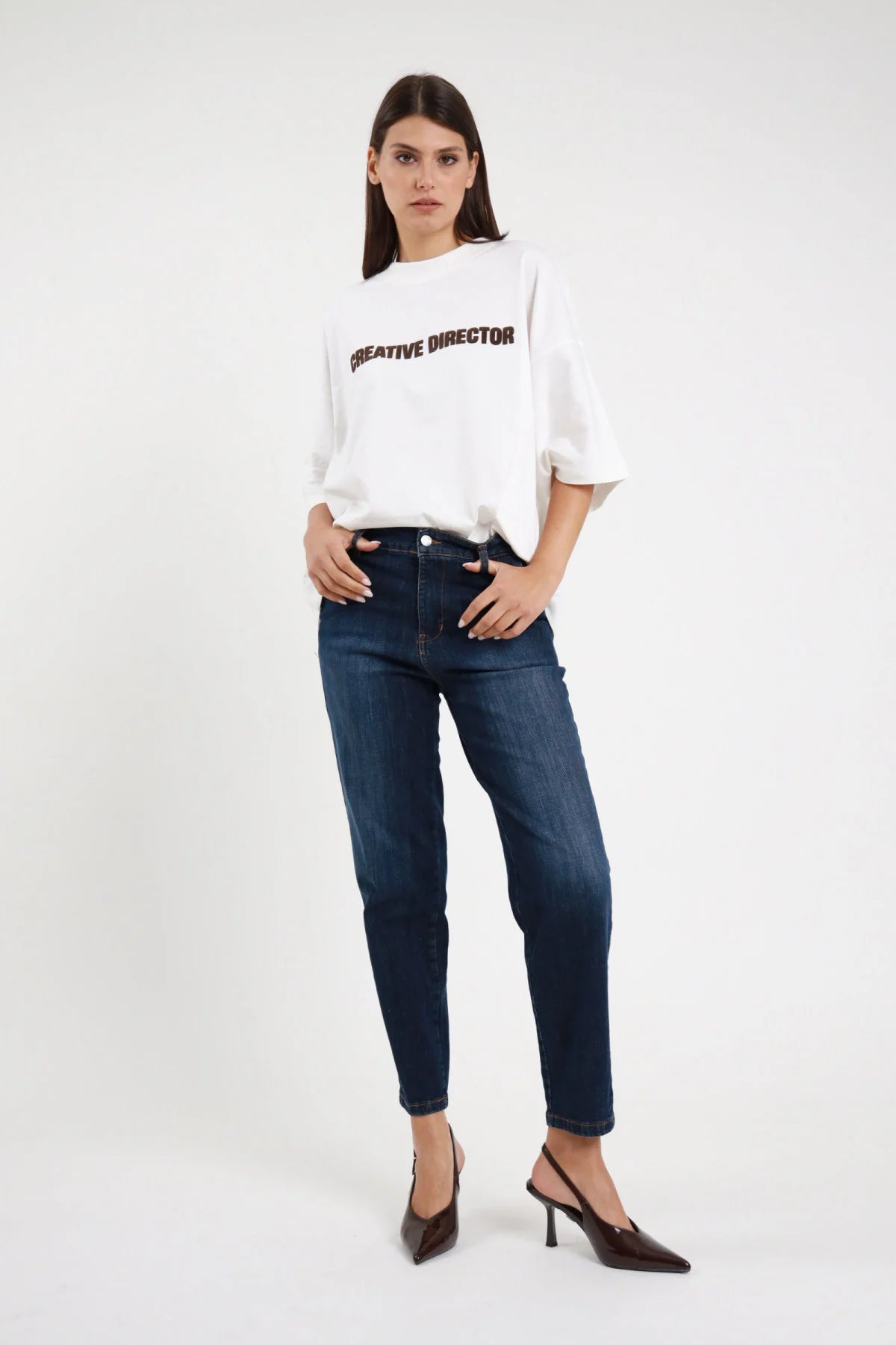 Jeans Chino Slim Vita Alta blu scuro indaco 98% cotone vita alta passanti cintura gamba dritta affusolata tasche frontali classiche bottone argento fondo cropped caviglia frontale intera - modella intera frontale posa statica mani jeans sguardo frontale intenso capelli castani lisci sfondo bianco t-shirt bianca Creative Director oversize slingback marrone tacco