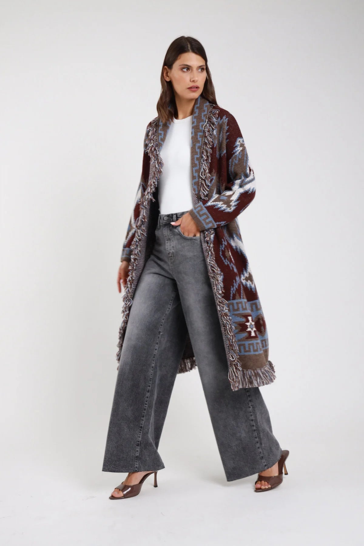 Jeans wide leg palazzo grigio stonewashed vita alta 5 tasche gamba larga maxi orlo grezzo frontale intera con cardigan azteco bordeaux frange - modella frontale intera sguardo laterale capelli castani scuri ondulati sfondo bianco cardigan azteco bordeaux frange t-shirt bianca sandali tacco