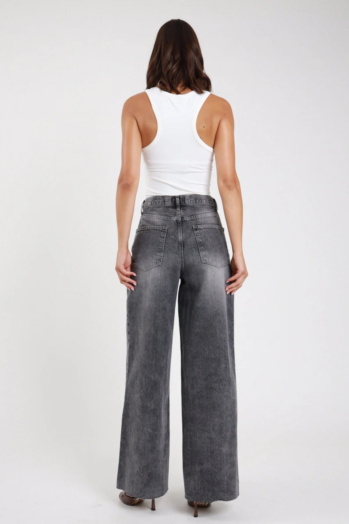 Jeans wide leg palazzo grigio stonewashed vita alta 5 tasche gamba larga maxi orlo grezzo retro intera - modella retro intera top bianco canotta sguardo non visibile capelli castani scuri ondulati sfondo bianco sandali tacco a spillo marrone