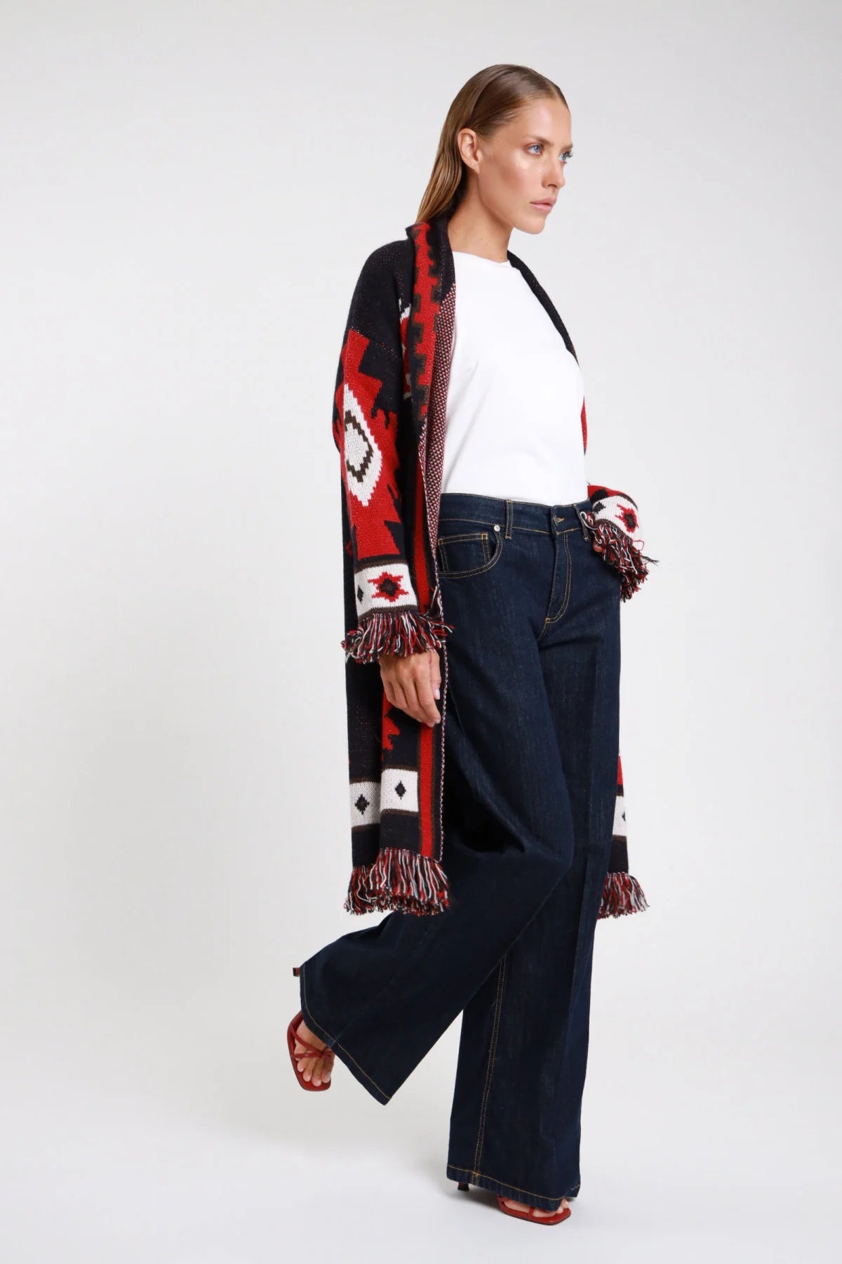 Jeans palazzo wide-leg blu indigo laterale in cammino silhouette full-length cardigan azteco frange - modella laterale intera in cammino t-shirt bianca cardigan azteco rosso nero frange sandali rossi capelli castani