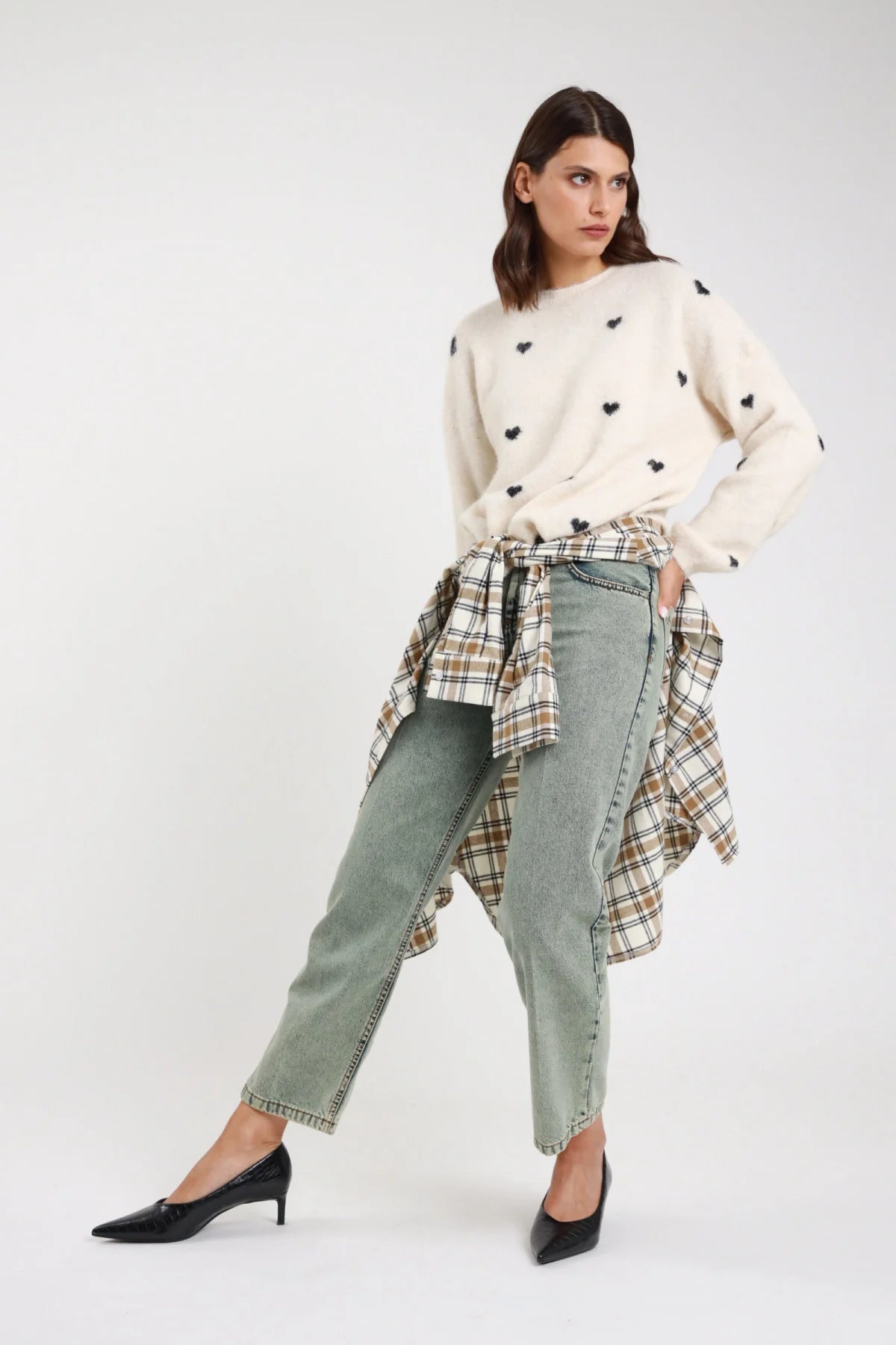 Jeans Manu straight leg verde sage acqua stonewashed rhinestone vita alta 5 tasche orlo grezzo frontale intera con pullover cuori avorio - modella frontale intera sguardo laterale capelli castani scuri mossi sfondo bianco pullover cuori avorio camicia check annodata vita décolleté neri kitten heel