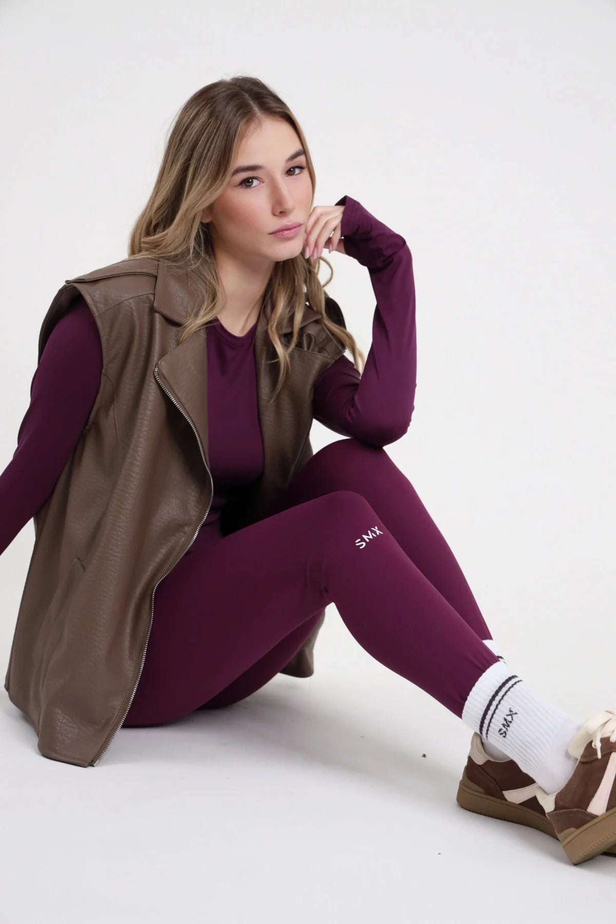 Leggings SMX Limited Edition viola melanzana logo SMX bianco gamba coscia laterale 95% tactel body line full length seduta 3/4 laterale mano mento - modella seduta 3/4 laterale mano mento sguardo morbido capelli biondi mossi sfondo bianco logo SMX gamba laterale visibile gilet biker pelle tortora aperto maglia second skin viola calzini SMX bianchi scarpe marrone abbinati