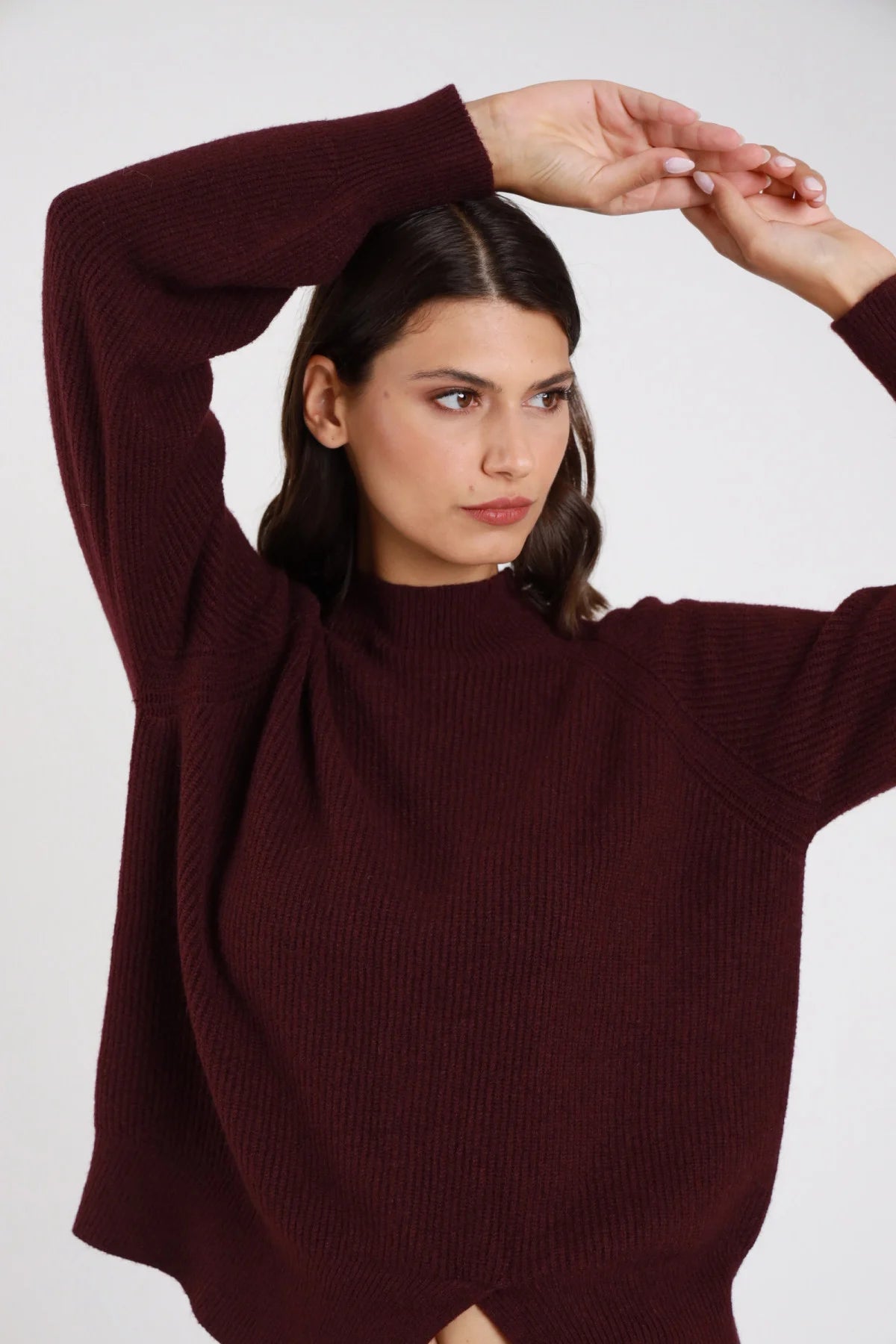 Pullover maglia coste mock neck collo semi-alto fondo asimmetrico spacco punta batwing oversize bordeaux vino frontale busto braccia alzate mani testa - modella frontale busto braccia alzate mani testa sguardo laterale capelli castani scuri lisci sfondo bianco