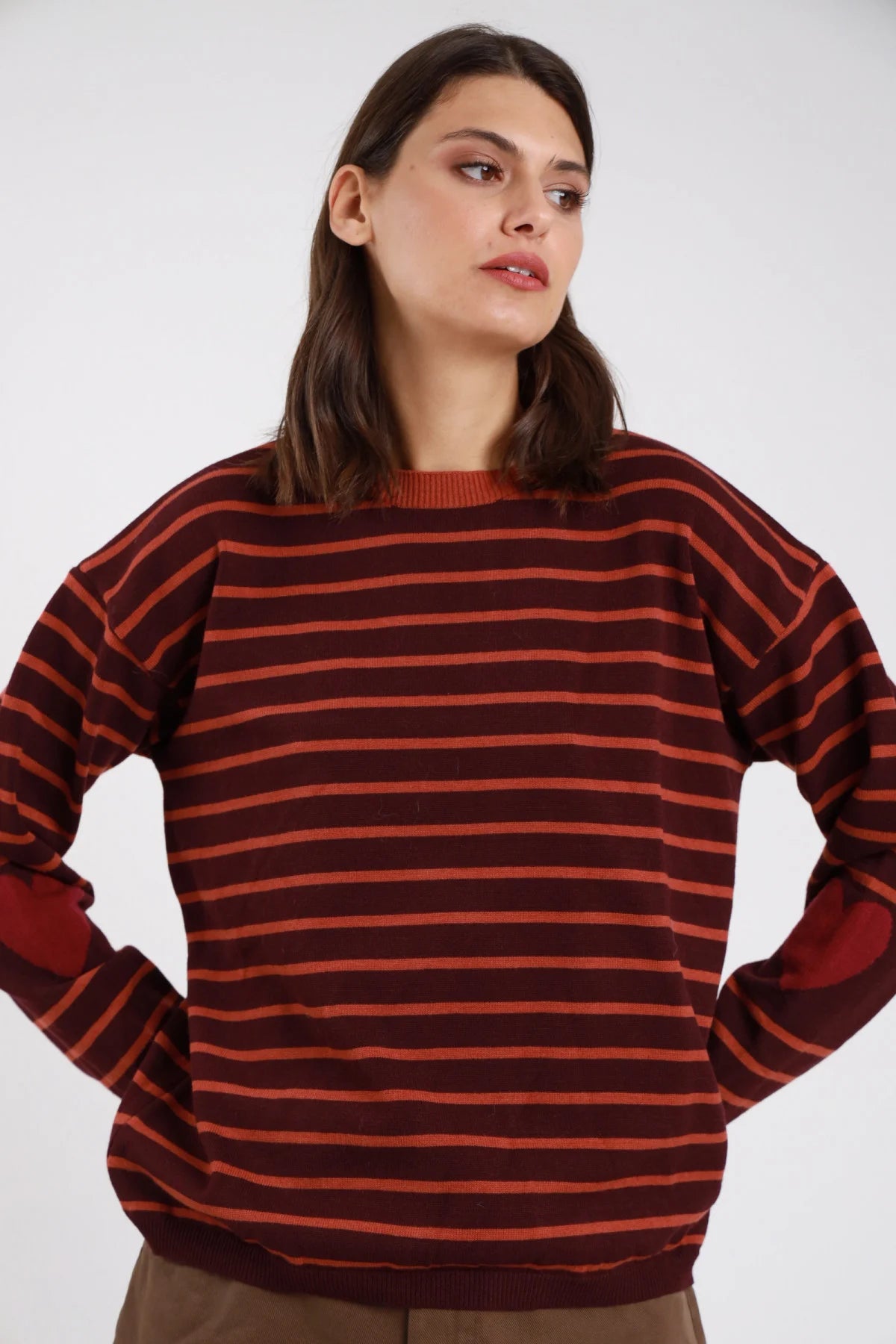 Pullover righe orizzontali bicolore cuori jacquard intarsiati gomiti bordeaux granata arancio ruggine cuori rosso bordeaux frontale mani fianchi - modella frontale mani fianchi sguardo laterale capelli castani scuri ondulati sfondo bianco pantaloni cammello cargo