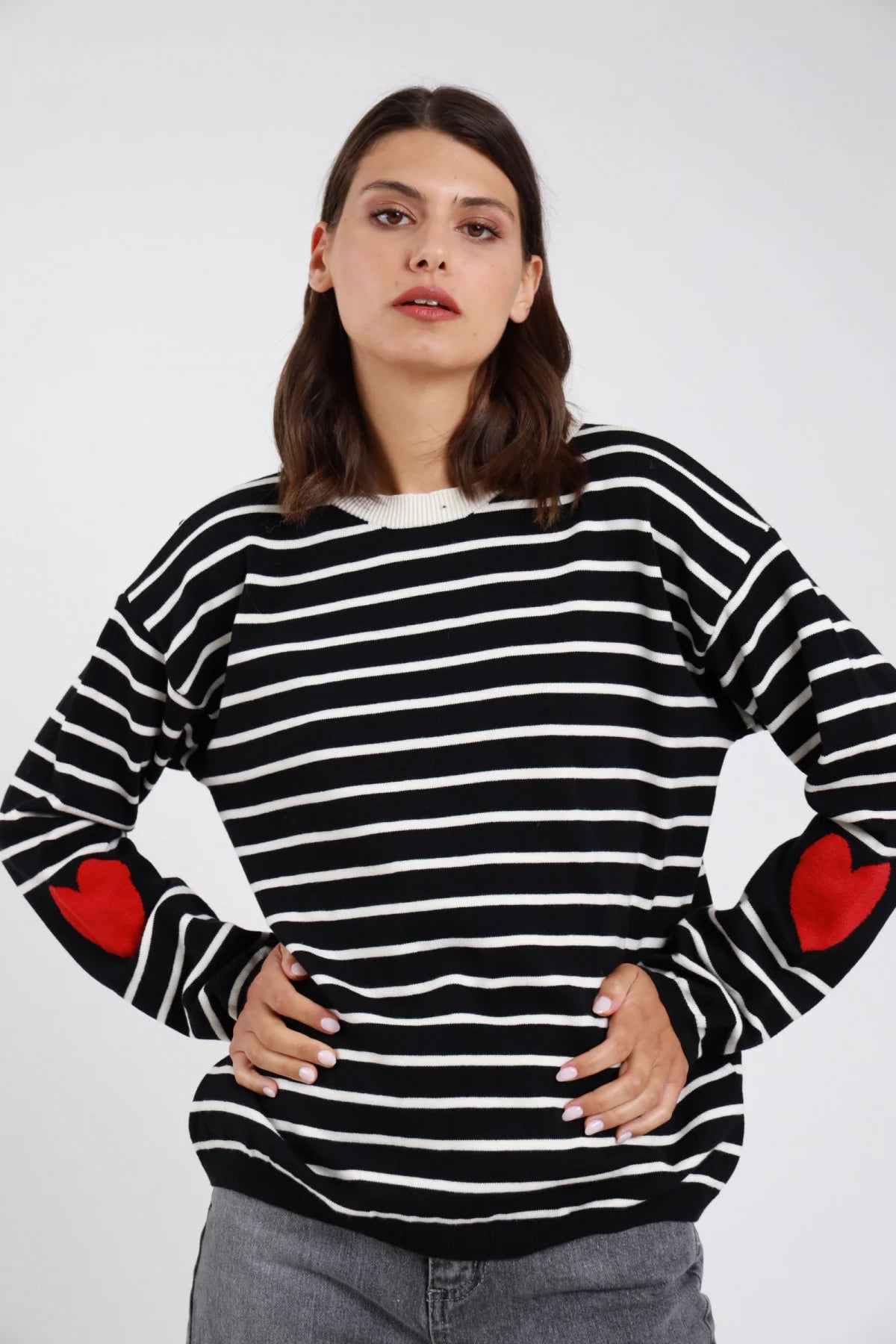 Pullover righe orizzontali bicolore cuori jacquard intarsiati gomiti nero bianco panna cuori rosso frontale mani fianchi - modella frontale mani fianchi sguardo frontale capelli castani scuri ondulati sfondo bianco jeans grigio scuro