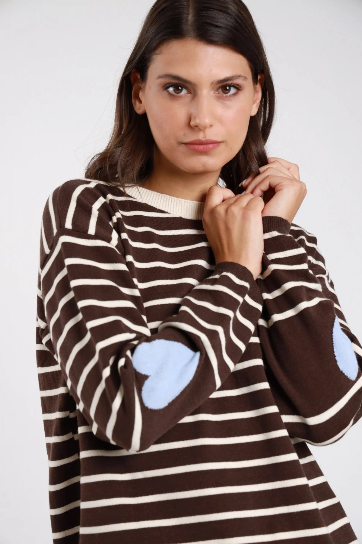 Pullover righe orizzontali bicolore cuori jacquard intarsiati gomiti marrone cioccolato avorio panna cuori azzurro baby blue close-up dettaglio cuore gomito mano collo - modella close-up busto mano collo sguardo laterale capelli castani scuri ondulati sfondo bianco dettaglio cuore azzurro gomito