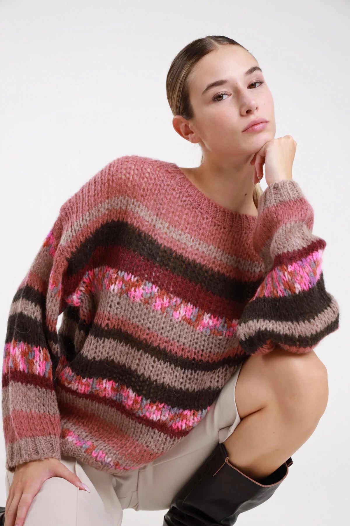 Maglione mohair righe rosa cammello bordeaux cioccolato jacquard astratto - modella busto tre quarti seduta mano al mento sguardo laterale pantalone beige stivali neri capelli castani raccolti