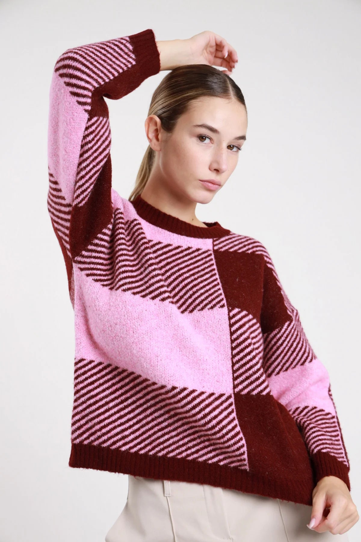 Pullover quadri patchwork bordeaux rosa cipria ciclamino frontale busto braccio alzato mano testa jacquard maxi check bicolor righe diagonali girocollo - modella frontale busto braccio alzato mano testa sguardo laterale basso pantaloni beige capelli castani raccolti