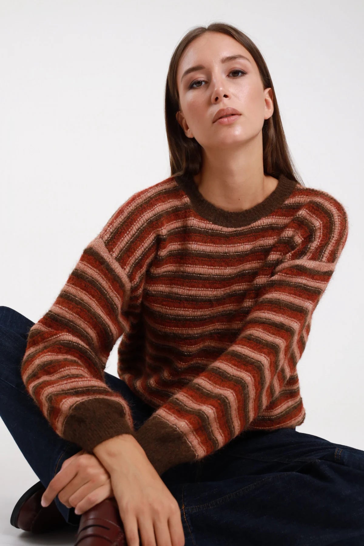 Maglione righe multicolor wave ruggine cotto cioccolato frontale seduta girocollo costina cioccolato manica balloon - modella frontale seduta sul pavimento gambe incrociate sguardo frontale jeans indaco stivali marroni capelli castani lisci