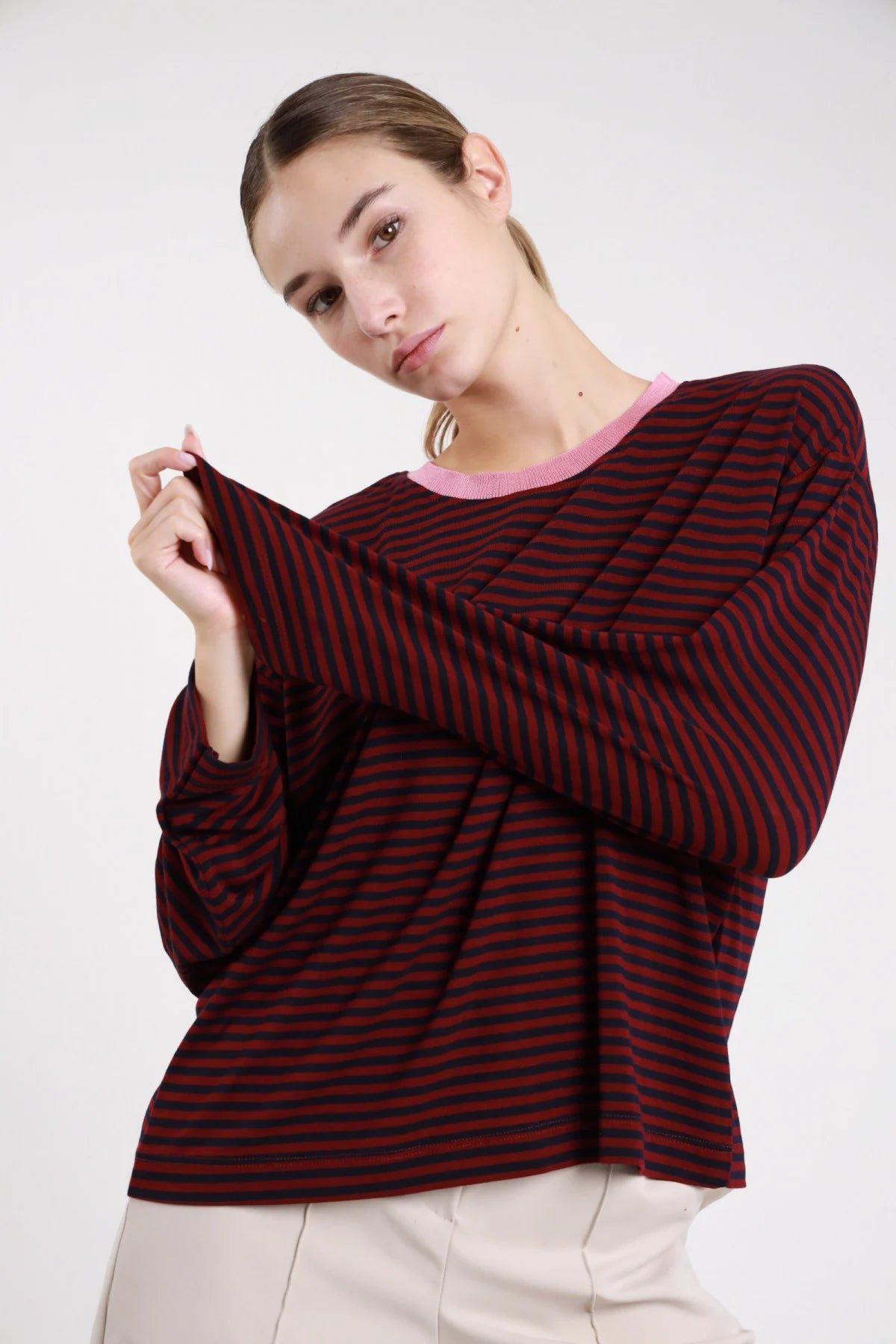 Maglia righe bicolor bordeaux navy frontale busto mano colletto girocollo costina rosa cipria manica lunga corpo oversize - modella frontale busto mano colletto sguardo laterale alto pantaloni beige capelli castani raccolti