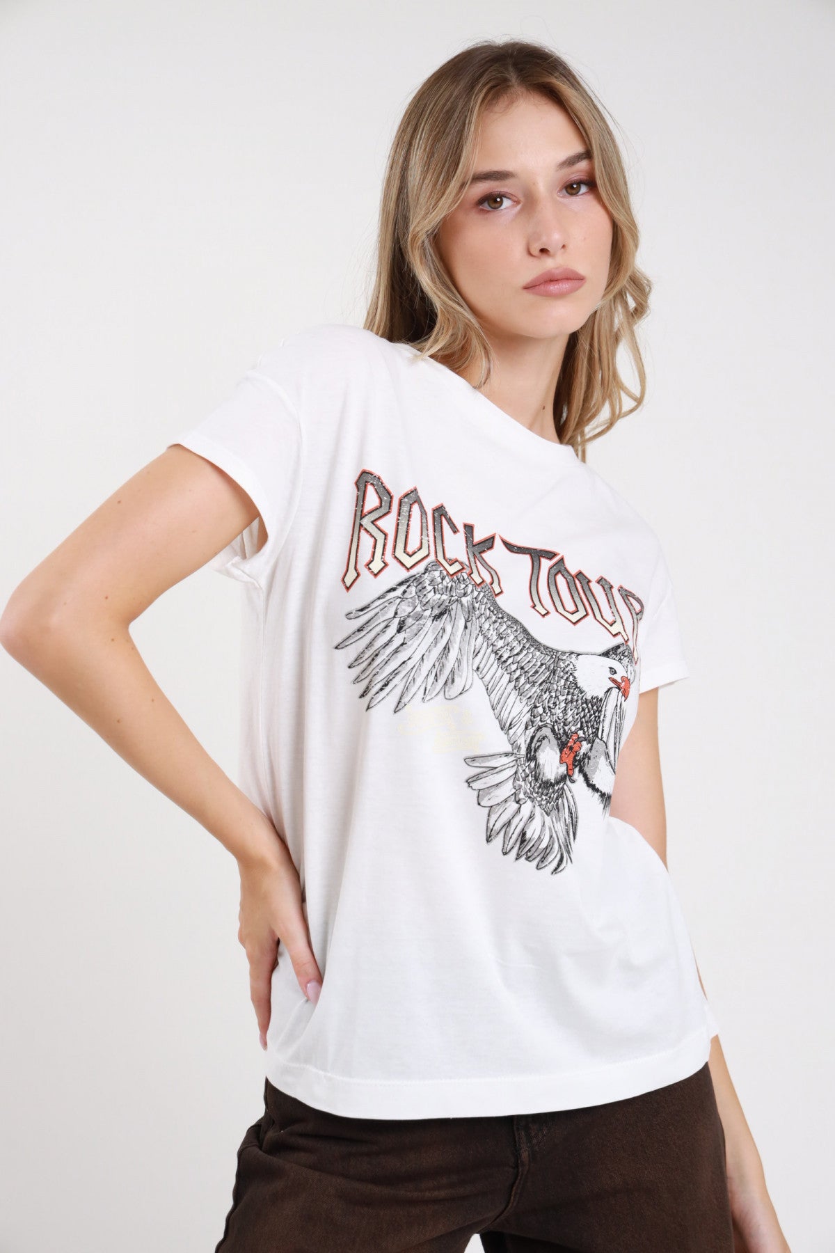 T-shirt rock tour grafica bianca busto tre quarti scollo V asimmetrico stampa aquila rock gotico - modella busto tre quarti mano fianco sguardo frontale pantaloni marrone scuro capelli biondi ondulati