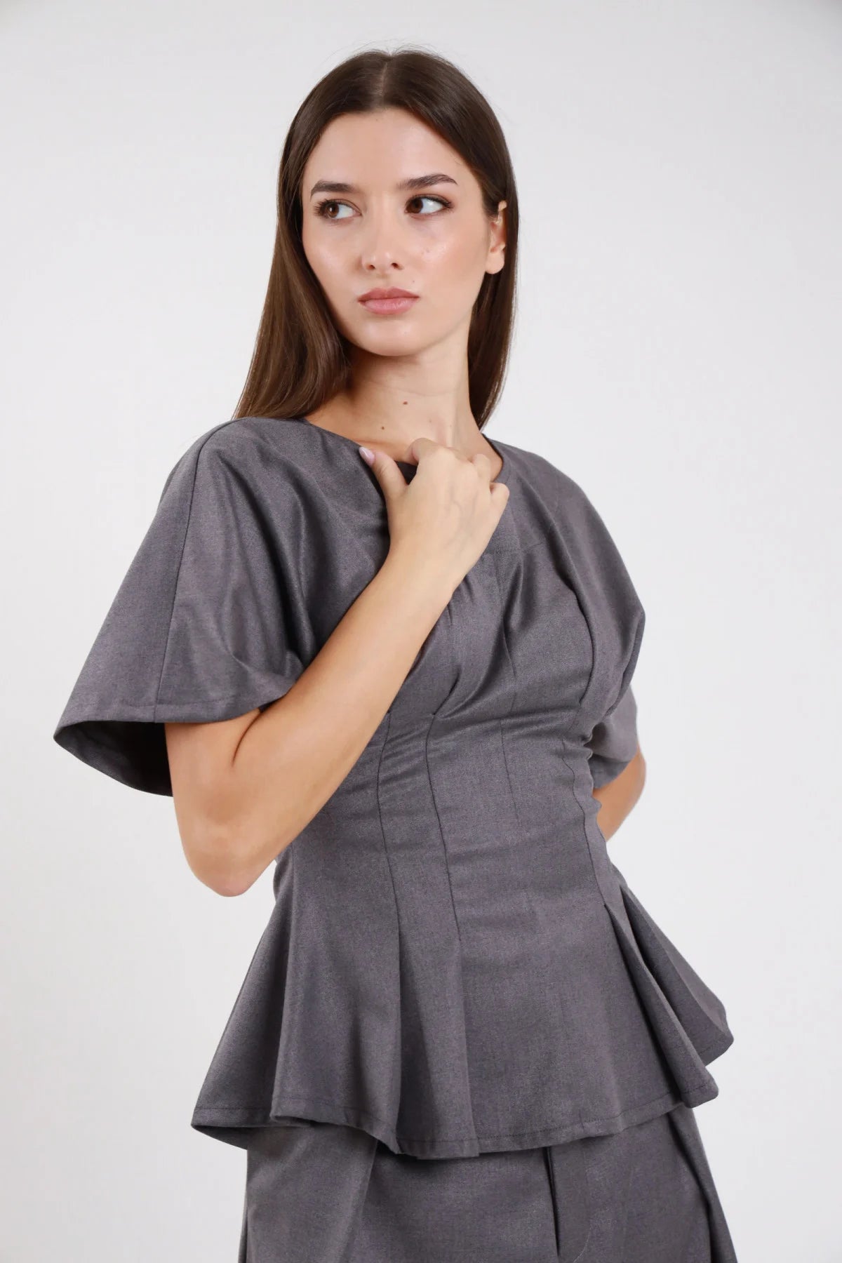 Top maglia sartoriale grigio ardesia antracite crêpe stretch corset pince verticali vita-definita manica corta flutter campana svasata peplum doppio balze sfalsate svasate girocollo tondo - modella laterale tre quarti mano collo sguardo laterale alto capelli castani scuri lisci sfondo bianco gonna check grigio a strati abbinata coordinata