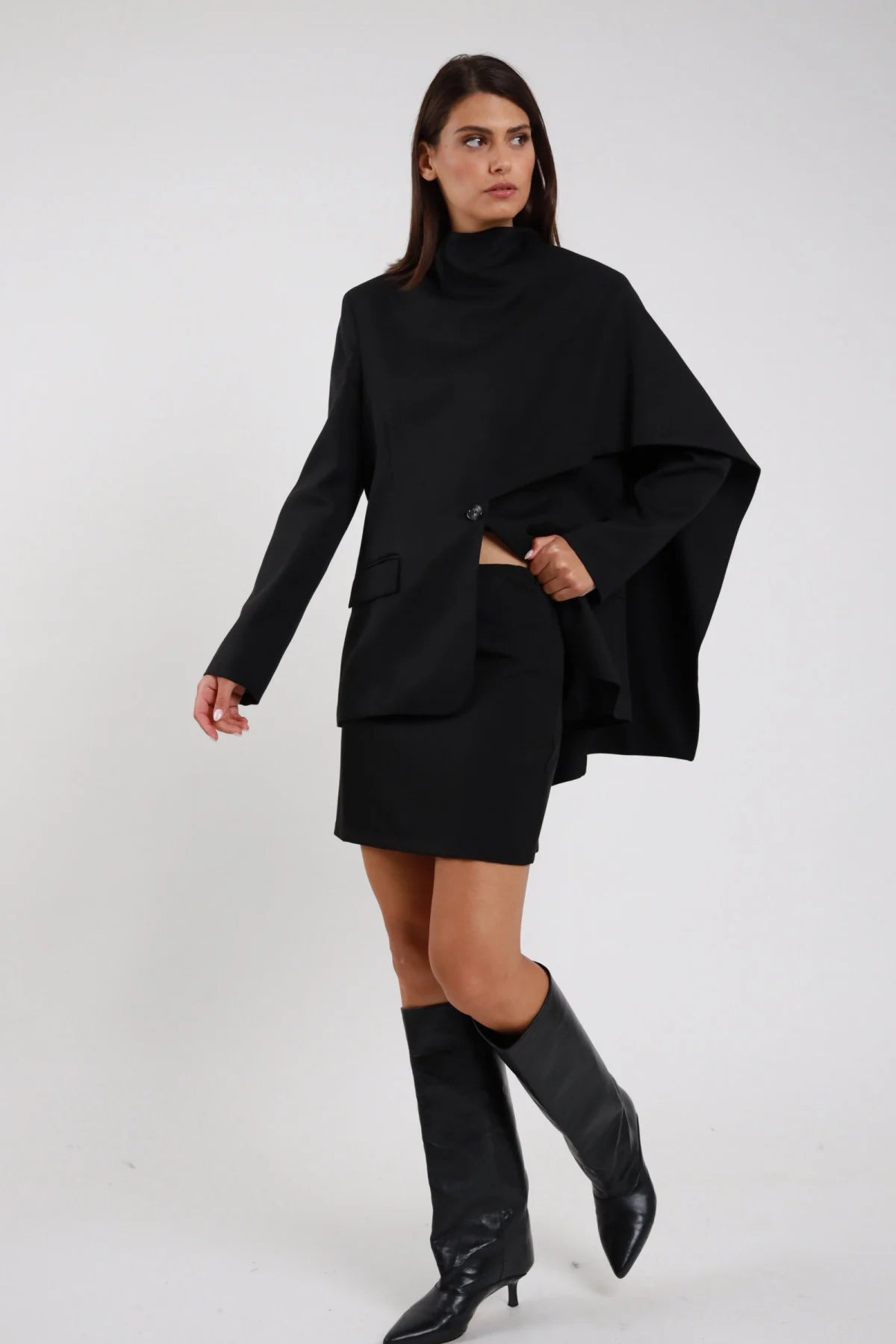 Minigonna sartoriale nero vita alta mini dritta tubino stretch frontale 3/4 posa camminata dinamica coordinata blazer mantella nero - modella frontale 3/4 posa camminata dinamica sguardo laterale capelli castani lisci sfondo bianco blazer mantella nero stivali neri alti ginocchio tacco kitten