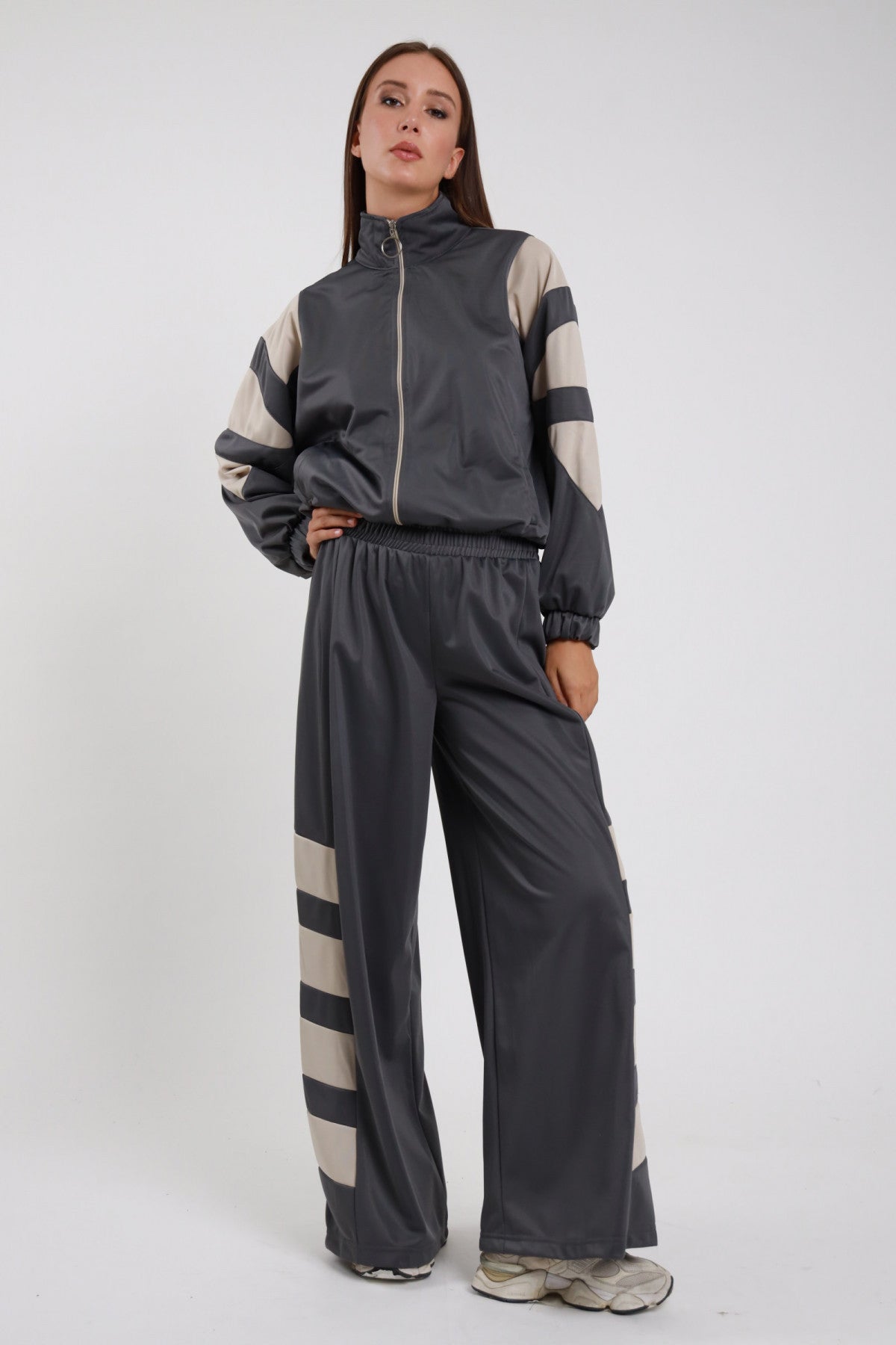 Pantaloni Palazzo Bande Diagonali antracite grigio scuro raso satin bande diagonali panna larghe obliquo vita alta elastico gamba wide leg palazzo caduta fluida frontale intera - modella intera frontale posa mano fianco sguardo frontale intenso capelli castani lisci sfondo bianco giacca zip antracite bande panna coordinata sneakers beige bianche
