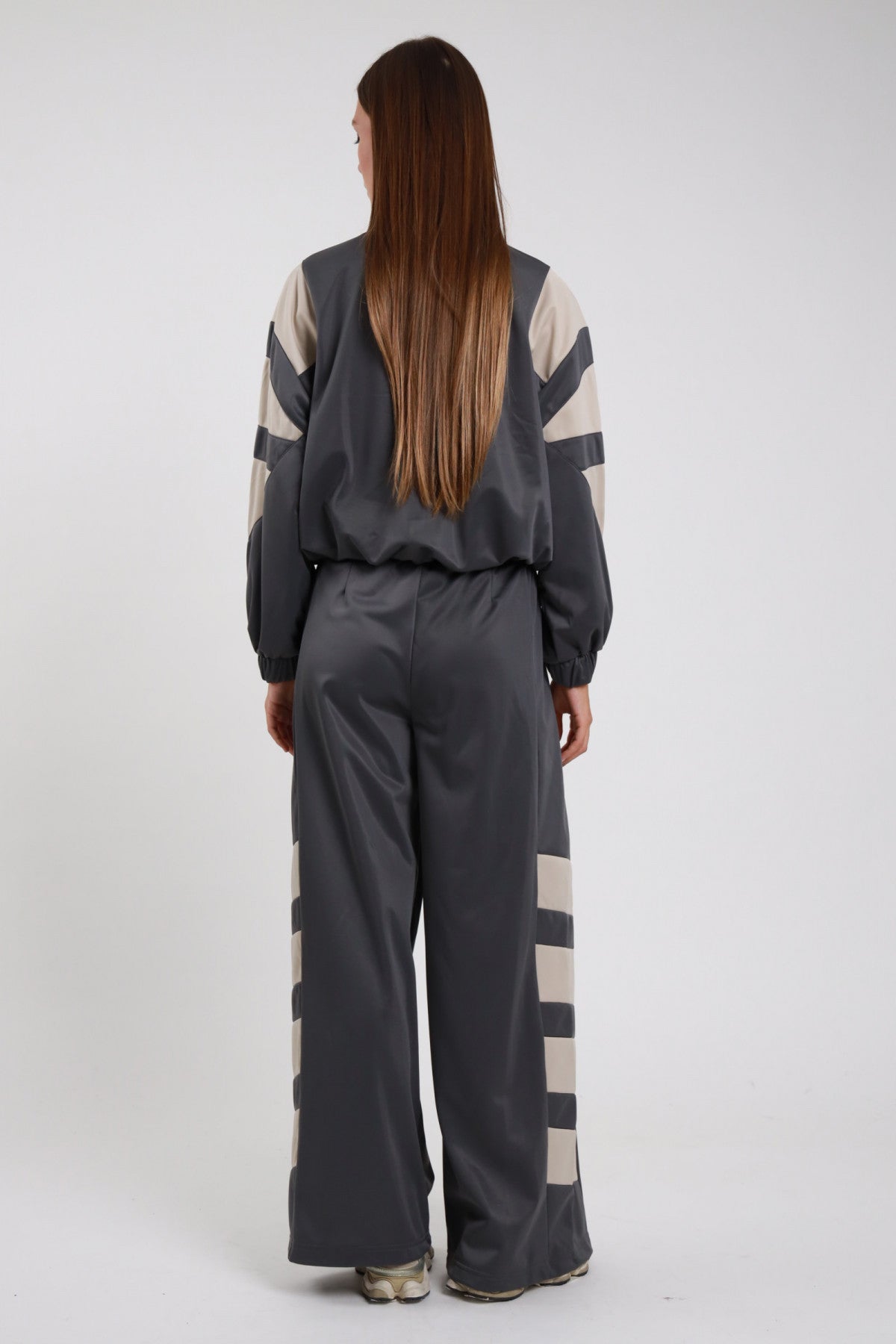 Pantaloni Palazzo Bande Diagonali antracite grigio scuro raso satin bande diagonali panna larghe obliquo vita alta elastico gamba wide leg palazzo caduta fluida posteriore intera - modella intera posteriore posa statica capelli castani lisci sfondo bianco giacca zip antracite bande panna coordinata sneakers beige bianche