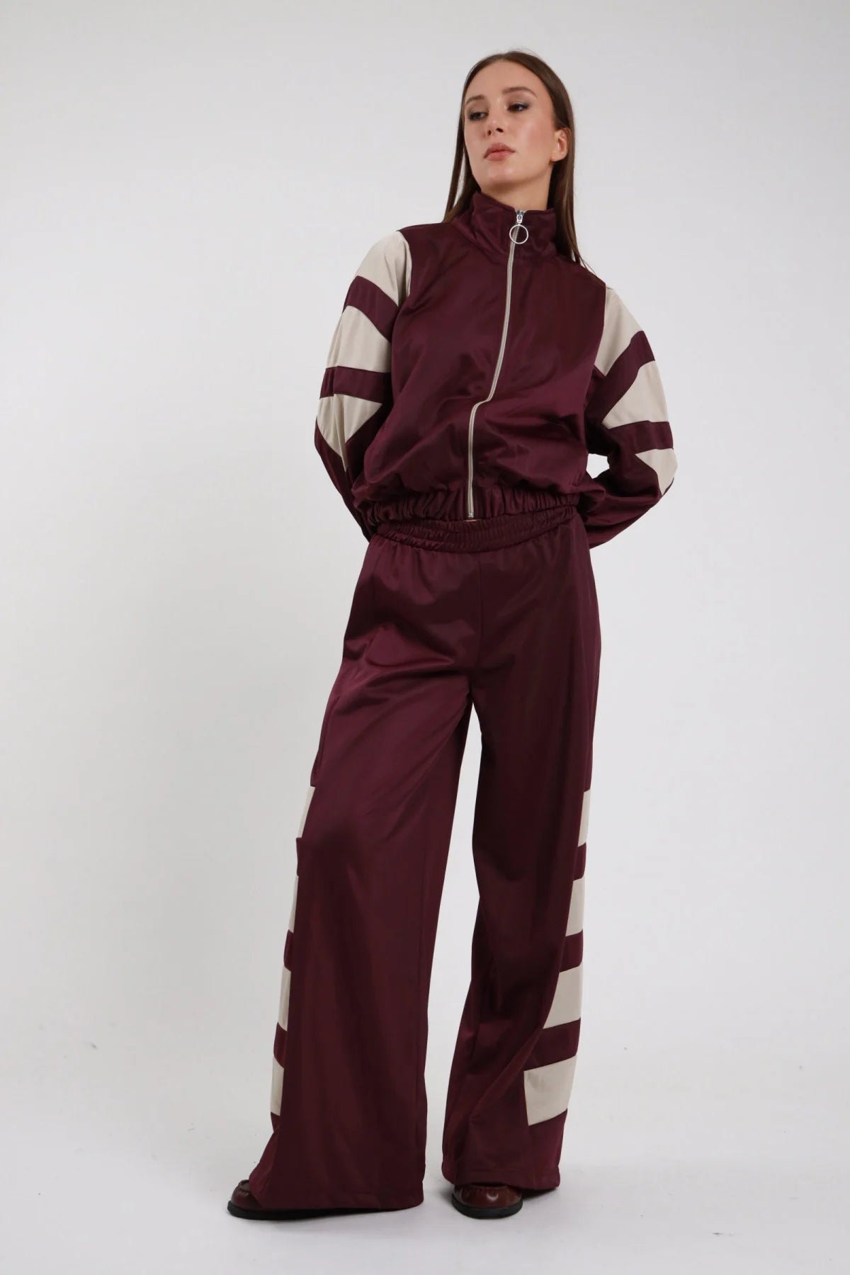 Pantaloni Palazzo Bande Diagonali bordeaux prugna raso satin bande diagonali panna larghe obliquo vita alta elastico gamba wide leg palazzo caduta fluida frontale intera - modella intera frontale posa mani tasche sguardo frontale intenso capelli castani lisci sfondo bianco giacca zip bordeaux bande panna coordinata scarpe marrone