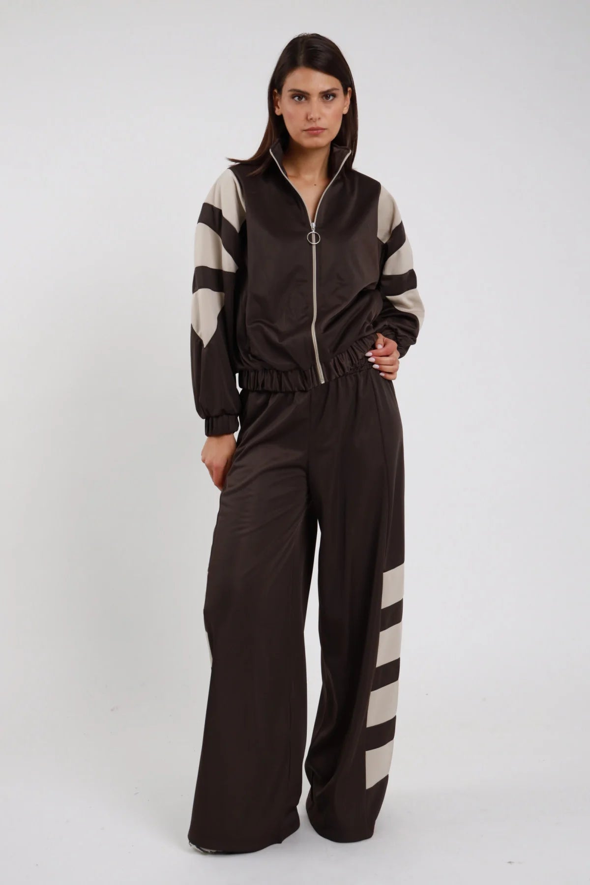 Pantaloni Palazzo Bande Diagonali marrone cioccolato scuro raso satin bande diagonali panna sabbia larghe obliquo vita alta elastico gamba wide leg palazzo caduta fluida frontale intera - modella intera frontale posa mano fianco sguardo frontale intenso capelli castani lisci sfondo bianco giacca zip marrone bande panna coordinata