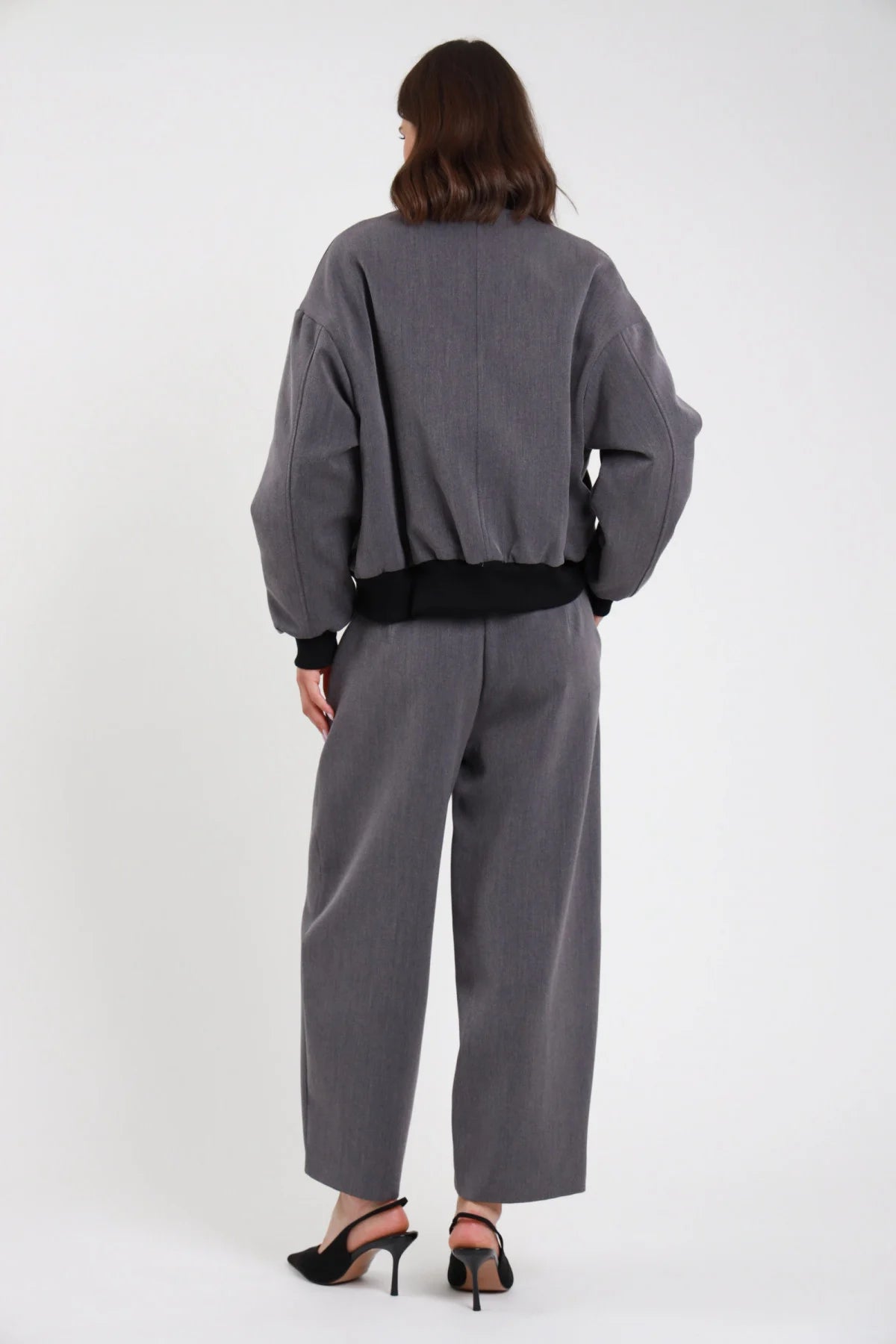 Pantalone Ovetto barrel grigio melange vita alta pinces tasche filetto gamba balloon scultorea cropped 7/8 retro intera con Bomber Alamaro grigio melange costine nere - modella retro intera sguardo non visibile capelli castani scuri sfondo bianco bomber grigio costine nere slingback neri tacco