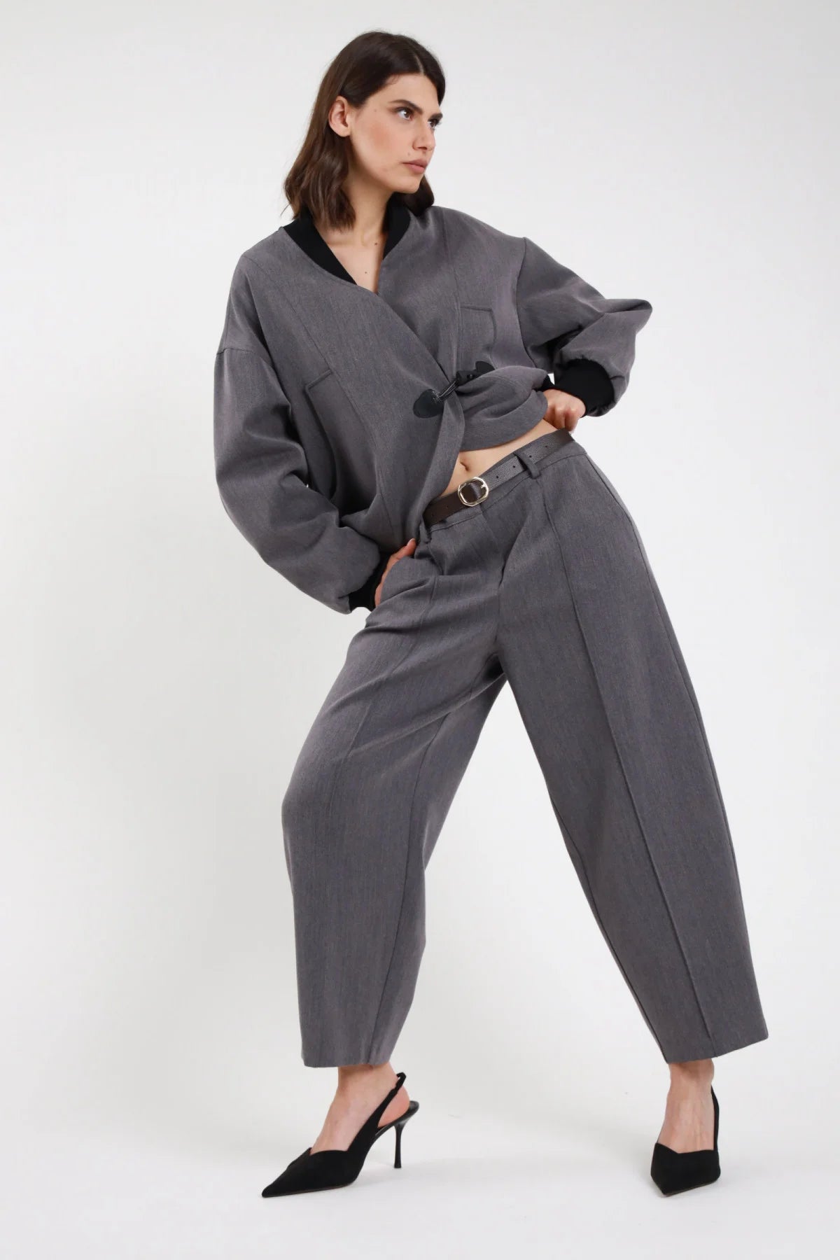 Pantalone Ovetto barrel grigio melange vita alta pinces tasche filetto gamba balloon scultorea cropped 7/8 frontale intera posa mossa con Bomber Alamaro e cintura thin dorata - modella frontale intera posa dinamica mossa sguardo laterale capelli castani scuri sfondo bianco bomber grigio cintura dorata slingback neri tacco