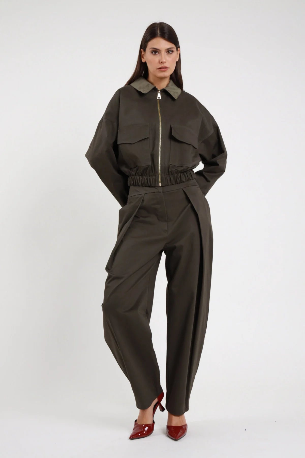 Pantalone piega Susy Mix Pre-Fall verde militare khaki 80% cotone 17% viscosa 3% elastan pince frontale strutturata vita alta gamba barrel ovetto tasche laterali fondo caviglia frontale intera mani tasche - modella intera frontale mani tasche sguardo frontale capelli scuri lisci sfondo bianco giacca crop zip stesso tessuto verde militare coordinata tacchi spillo bordeaux rosso abbinati