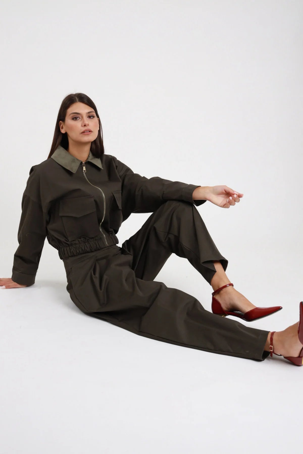 Pantalone piega Susy Mix Pre-Fall verde militare khaki pince frontale vita alta gamba barrel ovetto seduta 3/4 laterale - modella seduta pavimento 3/4 laterale sguardo laterale capelli scuri lisci sfondo bianco giacca crop zip stesso tessuto verde militare coordinata total look sartoriale tacchi spillo bordeaux rosso abbinati