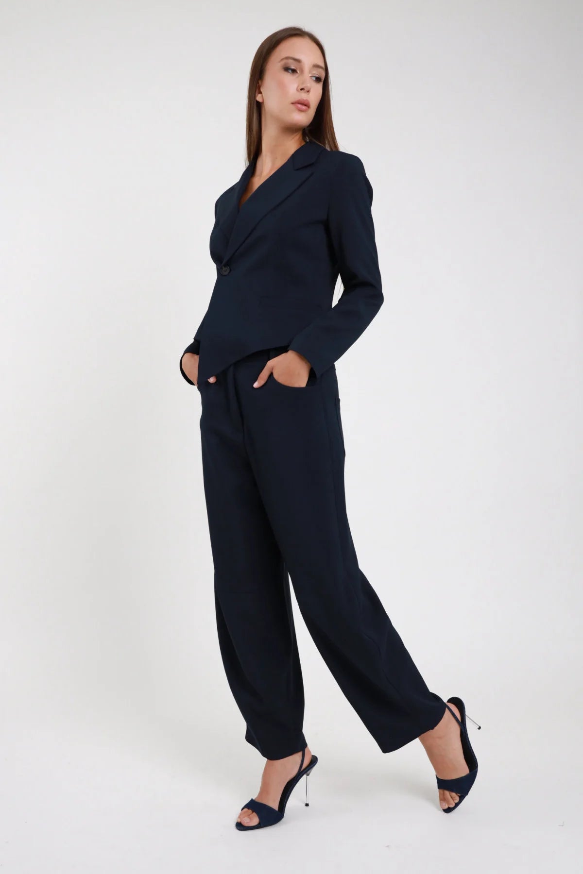 Pantalone Ovetto blu navy vita alta passanti cintura forma barrel scultorea pancia ampia orlo stretto caviglia intera laterale 3/4 posa dinamica mani tasche sguardo laterale - modella intera laterale 3/4 posa dinamica mani tasche sguardo laterale capelli castani sfondo bianco blazer asimmetrico navy coordinato sandali tacco spillo argento