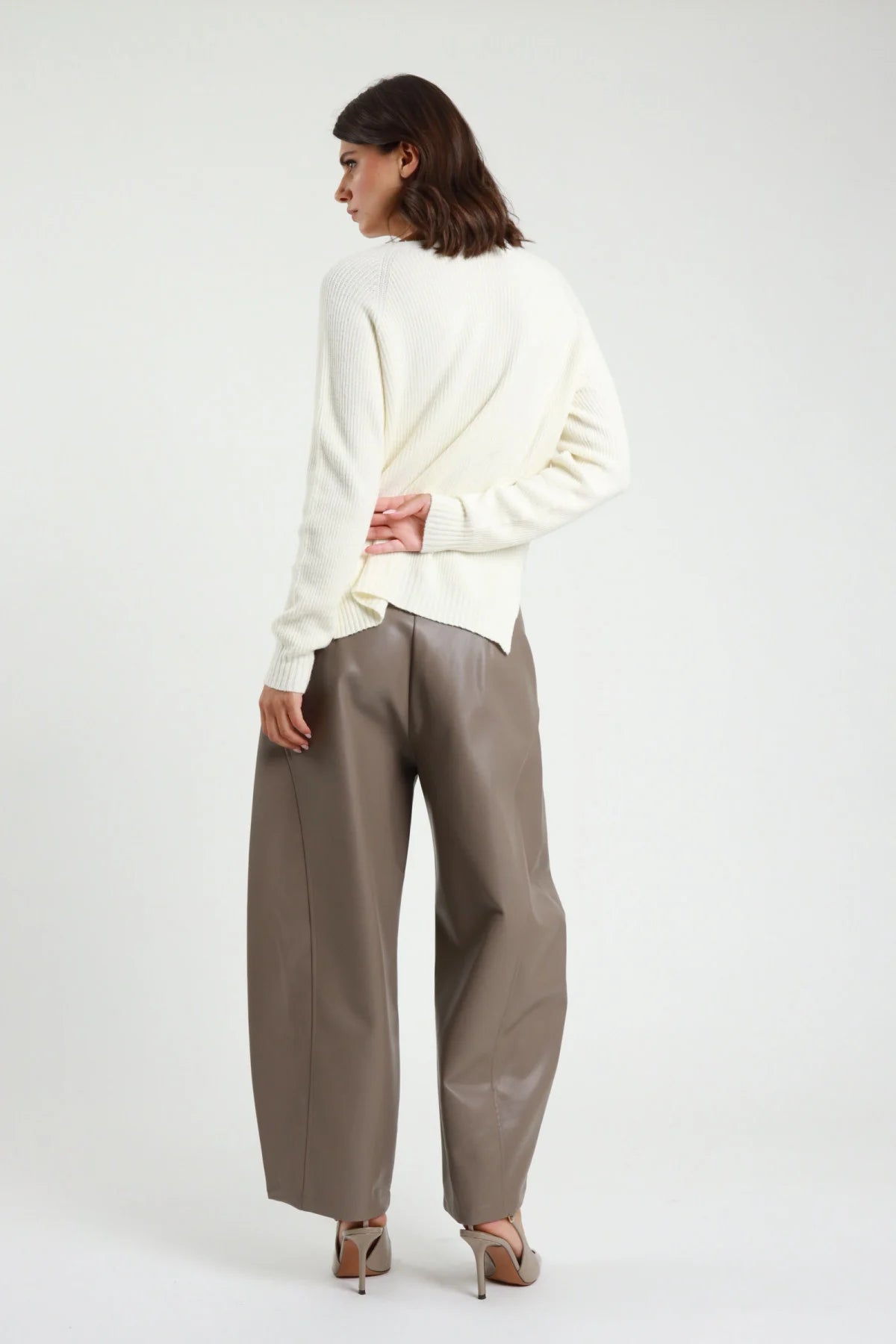 Pantalone Barrel Ecopelle tortora talpa vita alta pinces gamba barrel scultorea 7/8 cropped orlo dritto retro intera in piedi - modella retro intera mani schiena sguardo laterale capelli castani scuri sfondo bianco maglione bianco panna décolleté nude tacco