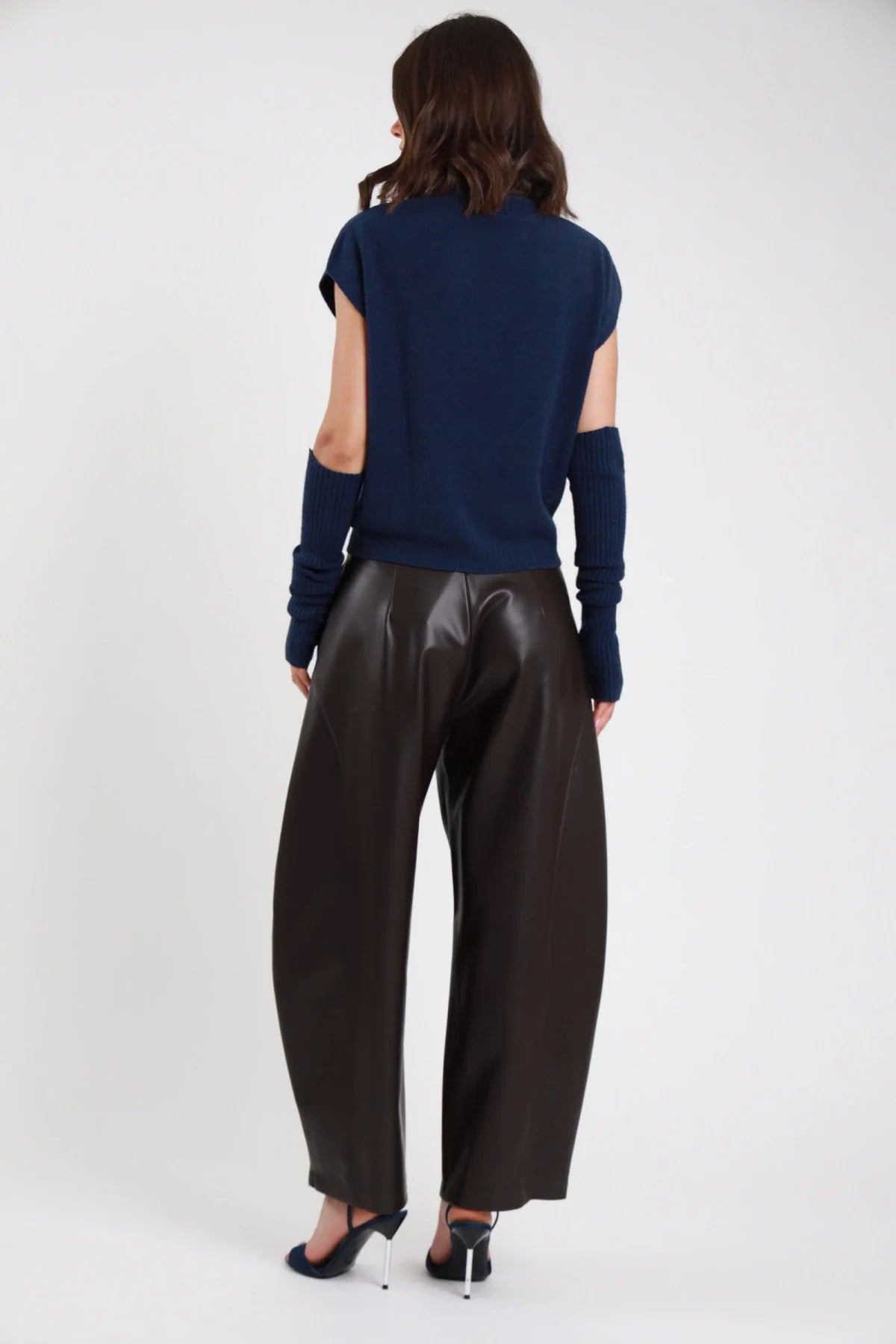 Pantalone Barrel Ecopelle marrone scuro cioccolato vita alta pinces gamba barrel scultorea 7/8 cropped orlo dritto retro intera in piedi - modella retro intera braccia lungo corpo sguardo laterale capelli castani scuri sfondo bianco top navy cut-out manica sandalo tacco argento