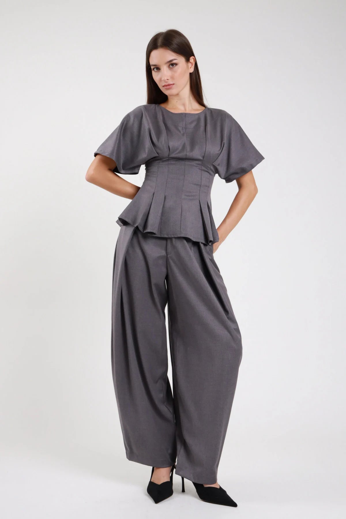 Pantaloni wide leg balloon grigio ardesia antracite crêpe stretch fascia elastica vita alta strutturata pince sartoriali drappeggio gamba ampia pouching volumetrico fianchi orlo dritto affusolato frontale intera - modella frontale intera mani fianchi sguardo frontale intenso capelli castani scuri lisci sfondo bianco top maglia zip corset peplum grigio coordinato décolleté nero tacco
