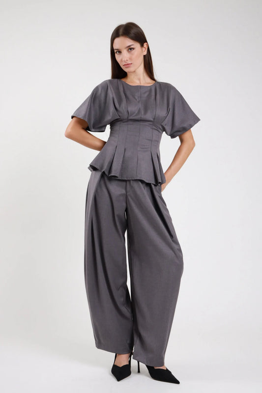 Pantaloni wide leg balloon grigio ardesia antracite crêpe stretch fascia elastica vita alta strutturata pince sartoriali drappeggio gamba ampia pouching volumetrico fianchi orlo dritto affusolato frontale intera - modella frontale intera mani fianchi sguardo frontale intenso capelli castani scuri lisci sfondo bianco top maglia zip corset peplum grigio coordinato décolleté nero tacco