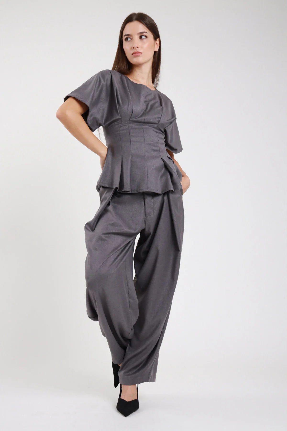 Pantaloni wide leg balloon grigio ardesia antracite crêpe stretch fascia elastica vita alta pince drappeggio gamba ampia svasata pouching volumetrico fianchi laterale dinamica - modella laterale tre quarti mano fianco sguardo laterale alto capelli castani scuri lisci sfondo bianco top maglia zip corset peplum grigio coordinato décolleté nero tacco