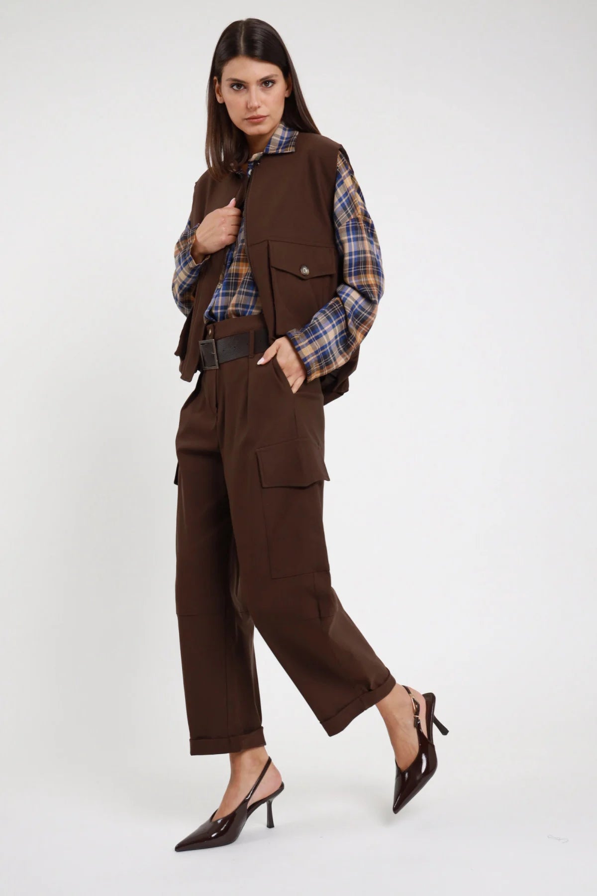 Pantalone Cargo Tasconi Vita Alta marrone cioccolato 78% viscosa pieghe davanti tasconi cargo gamba barrel ballon risvolto fondo cropped 3/4 laterale camminata - modella intera 3/4 laterale posa camminata dinamica capelli castani lisci sfondo bianco gilet marrone coordinato camicia quadri tartan slingback vernice marrone